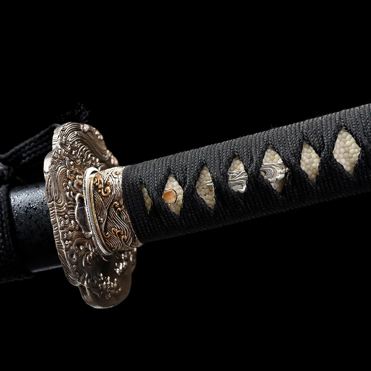 Détail rapproché du manche tsuka du katana japonais artisanal Kazekiri, tressage ito noir traditionnel laissant apparaître la peau de raie blanche samegawa avec motifs losangés réguliers, présence des mekugi en bambou, garde tsuba finement sculptée en métal aux motifs ondulés, finitions dorées et argentées visibles, fond noir profond mettant en valeur les textures e

