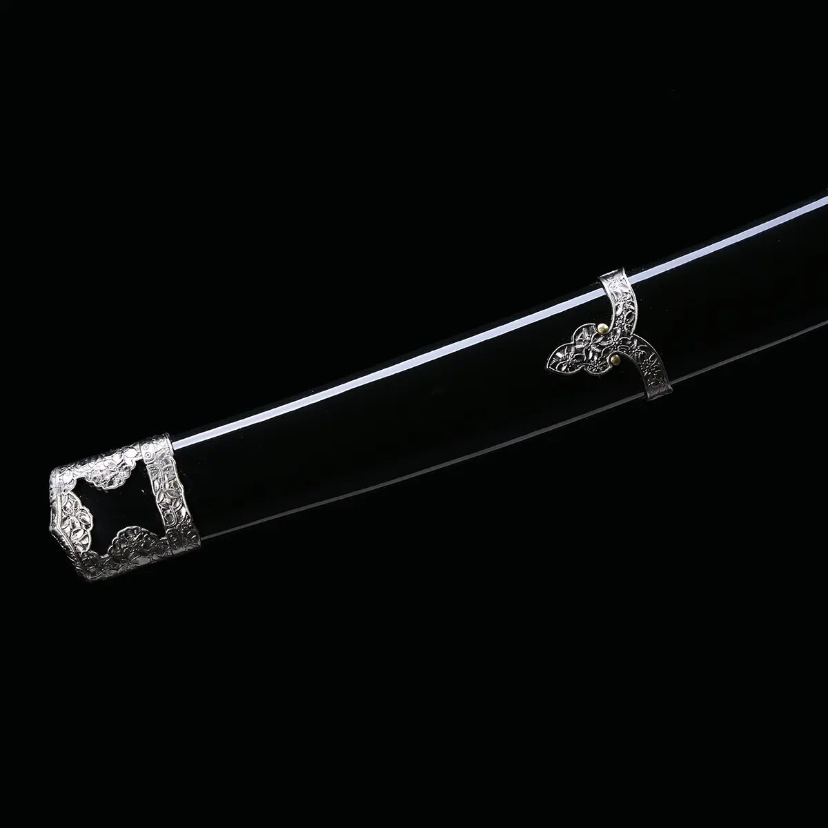 Détail du fourreau du katana japonais artisanal Seiryū no Tachi photographié sur fond noir uni. Le saya noir laqué est visible avec ses garnitures métalliques décoratives sculptées, incluant la kojiri et les attaches traditionnelles. La surface du fourreau présente une finition lisse et brillante, tandis que les éléments métalliques argentés montrent des motifs floraux finement gravés le long de la courbure.
