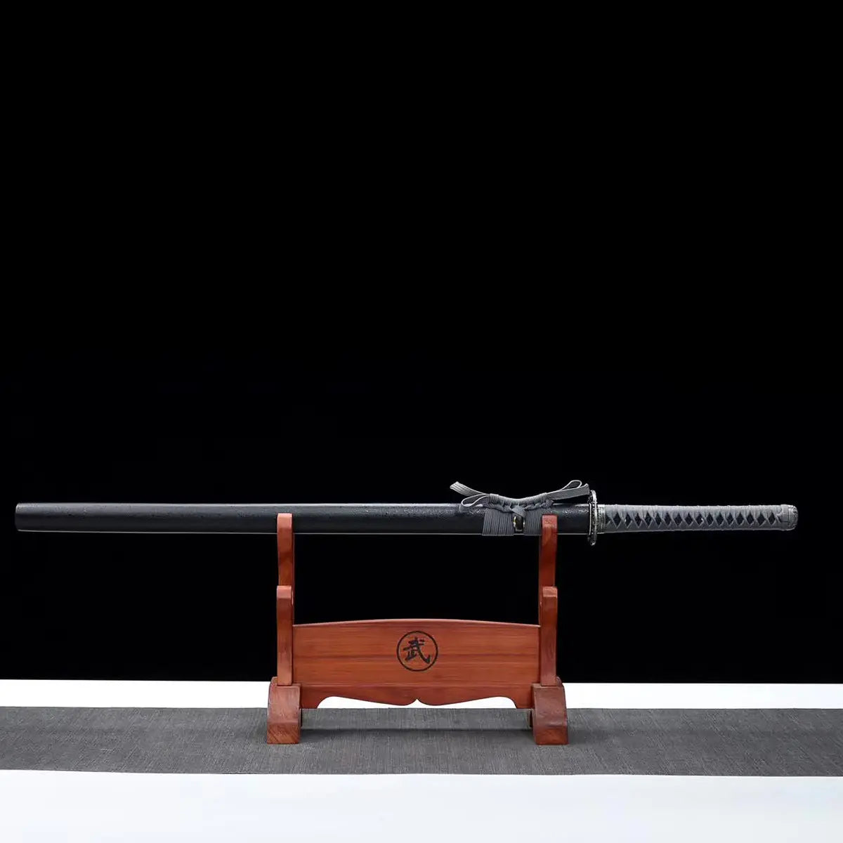 Katana japonais ONI NO KIBA dans son fourreau noir laqué posé horizontalement sur un support en bois rouge, poignée gris tressé avec sageo assorti, tsuba noire sculptée, fond noir avec surface grise.
