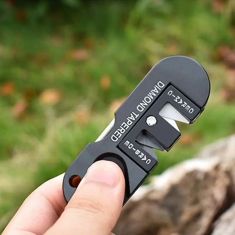 Aiguiseur de poche multifonction Blade Bushido tenu en main en extérieur, photographié devant un fond vert naturel avec herbe et pierre, mettant en valeur son design noir marqué “Diamond Tapered”, outil compact idéal pour l’affûtage des couteaux de survie et de camping.