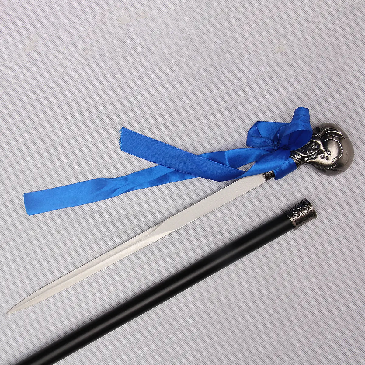 Vue rapprochée de la canne-épée “Blue Noble Cane” inspirée de Ciel Phantomhive : la lame inox polie est entièrement sortie, posée sur un fond gris clair texturé, juste au-dessus du tube en acier laqué noir. La poignée sculptée métallique argentée et son long ruban bleu contrastent avec le fond, mettant en valeur les détails du métal et la finesse de la lame.