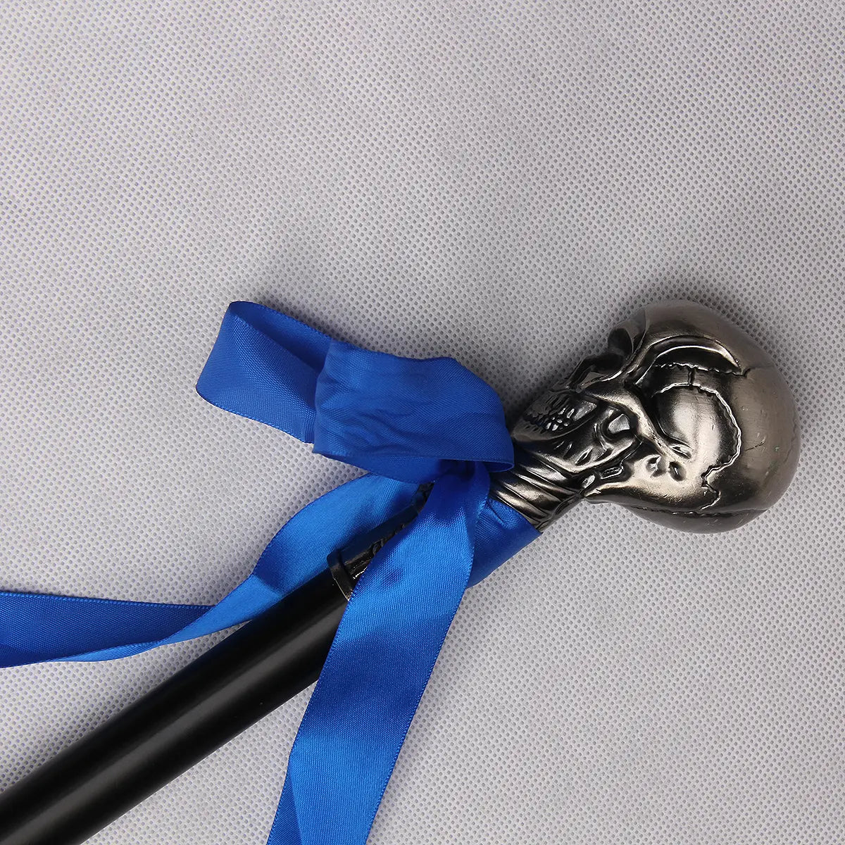 Gros plan de la poignée de la canne-épée “Blue Noble Cane”, tête sculptée en forme de crâne métallique argenté avec détails gravés, entourée d’un ruban bleu vif. La canne est posée sur un fond gris clair texturé, mettant en valeur le contraste entre le métal poli, le tissu et le tube acier laqué noir.