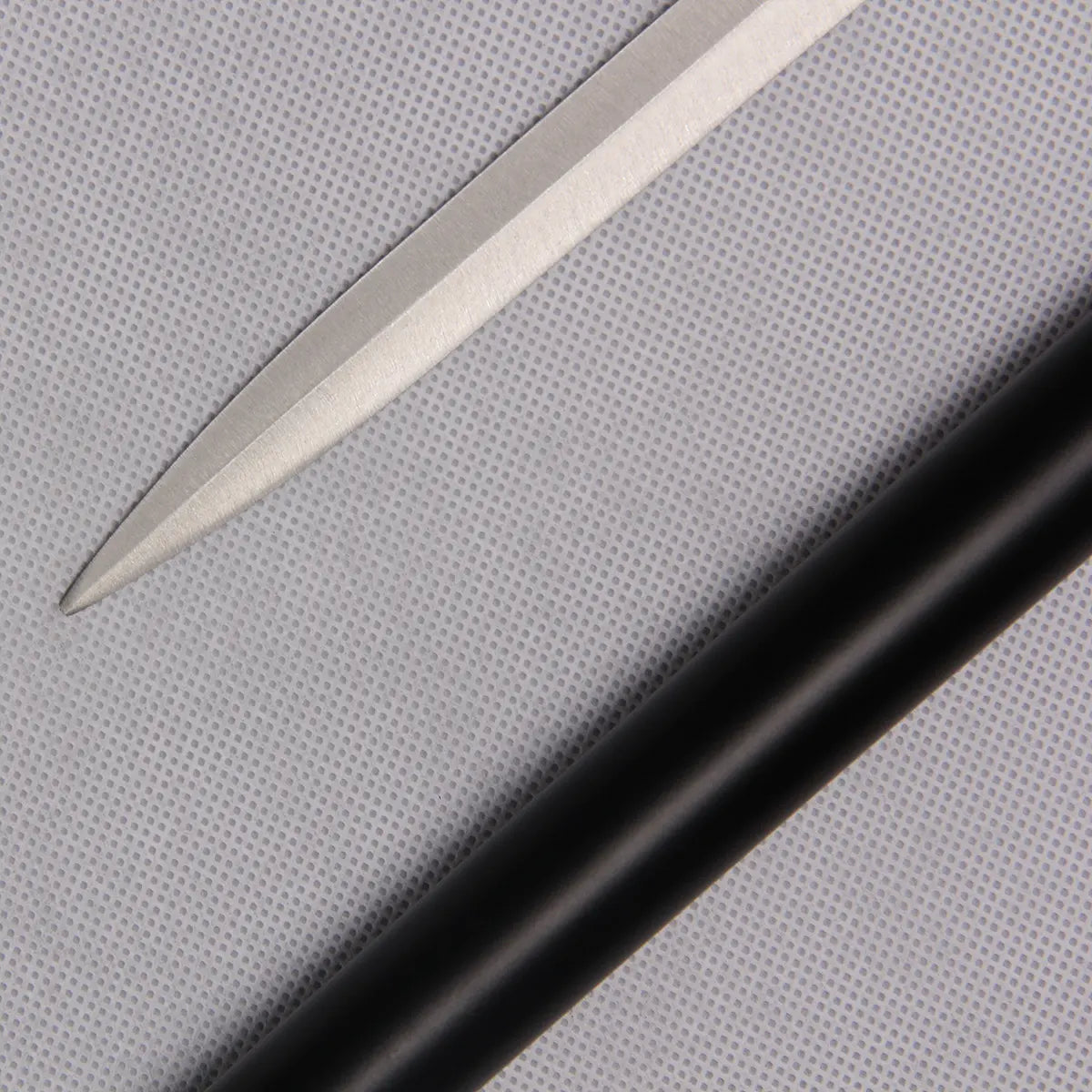 Gros plan de la pointe de la lame inox de la canne-épée “Blue Noble Cane”, posée sur un fond gris clair texturé. Le tube en acier laqué noir est visible en parallèle, mettant en valeur le contraste entre le métal poli et la finition mate du fourreau. Image centrée sur les détails de la lame et la qualité du polissage.