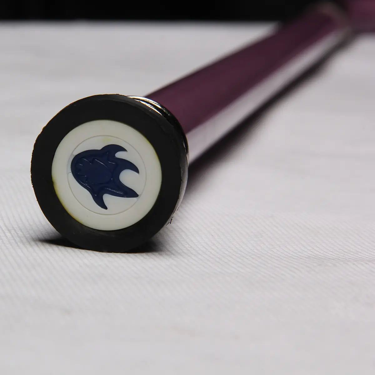 Gros plan de l’embout de la canne-épée “Dead Bone Blade”, posé sur un fond gris clair texturé ; on distingue le symbole bleu incrusté dans la base circulaire, avec le tube en acier laqué violet légèrement flou en profondeur, éclairage studio mettant en valeur les détails du motif et la brillance de la laque.