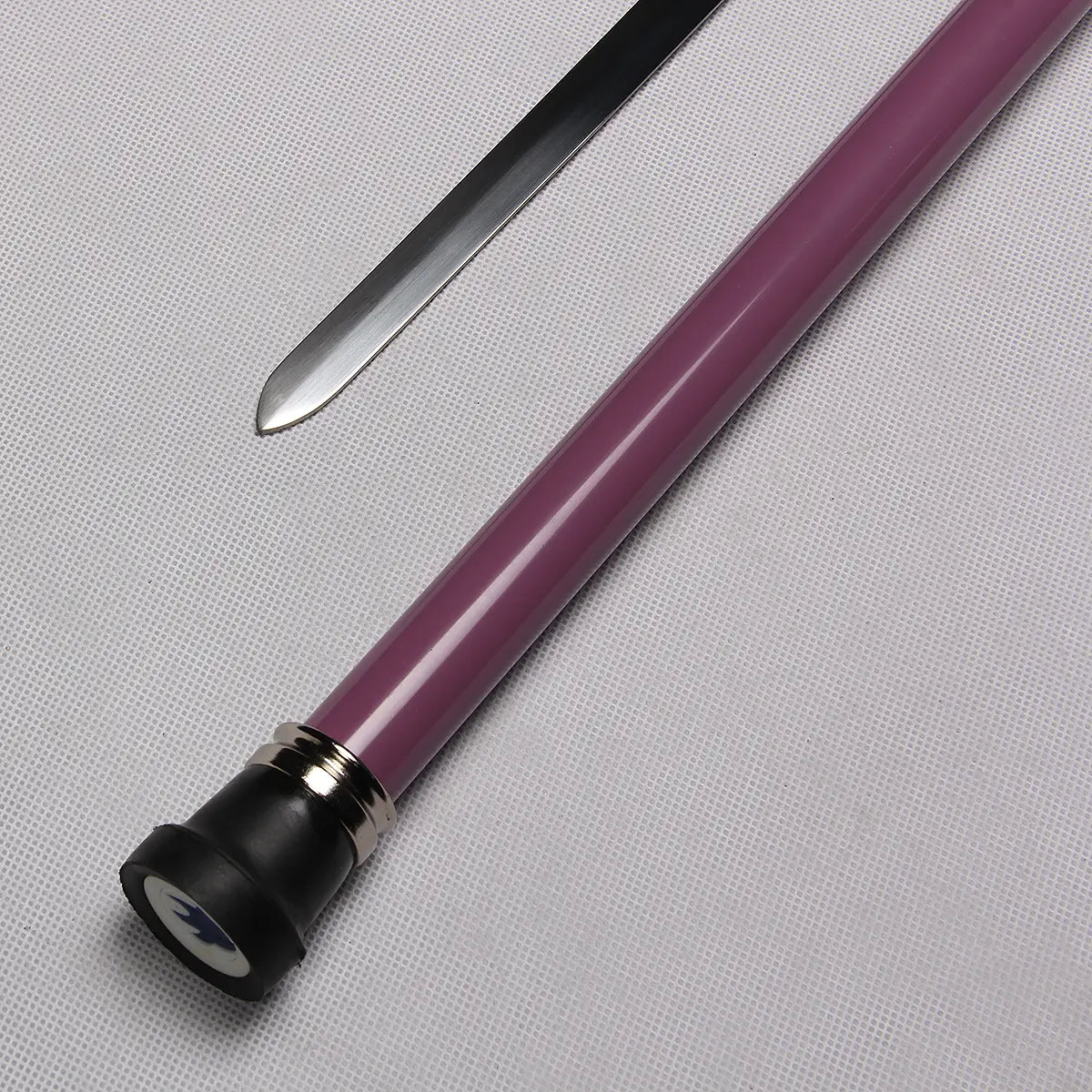 Gros plan de la canne-épée “Dead Bone Blade” montrant la pointe émoussée de la lame inox polie et le tube en acier laqué violet, posés sur un fond gris clair texturé ; l’embout noir avec bague métallique est visible au bas du fourreau, éclairage studio révélant les reflets du métal et la brillance de la laque.