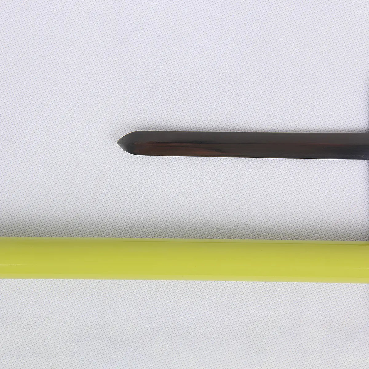 Gros plan de la pointe émoussée de la lame métallique d’une canne-épée cosplay couleur gris foncé, posée sur un fond textile blanc texturé, avec en bas une partie du fourreau tubulaire laqué vert clair.