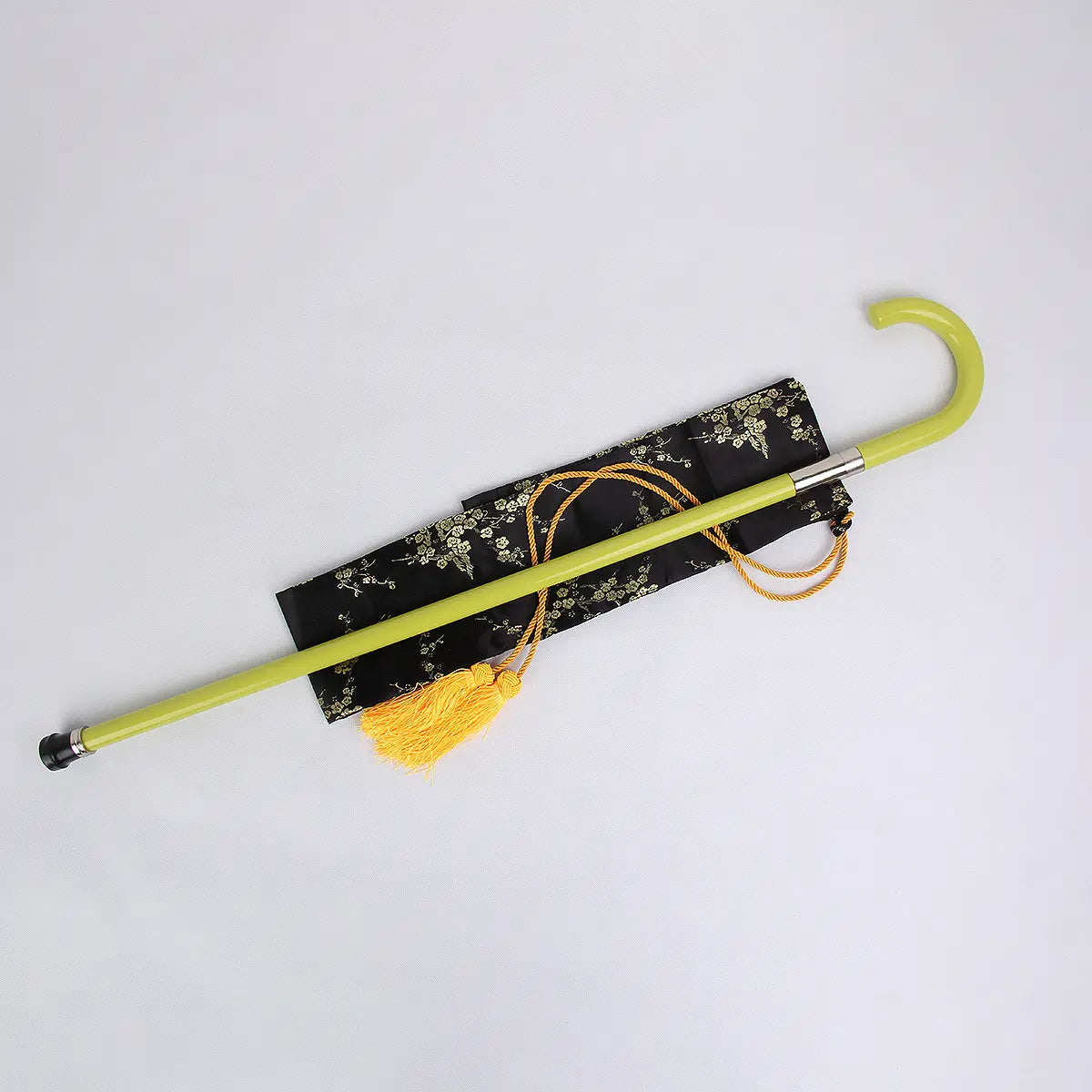 Canne-épée cosplay jaune inspirée de Kisuke Urahara posée sur une housse noire à motifs floraux dorés, fond gris clair uniforme, cordon décoratif jaune attaché, crochet arrondi et bague métallique clairement visibles.