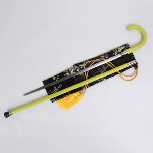 Gros plan d’une canne-épée cosplay inspirée de Kisuke Urahara (Bleach), posée sur une housse noire décorée de motifs floraux dorés. Le tube laqué vert clair est ouvert, laissant apparaître la lame fine en acier inoxydable ; un pompon jaune est attaché à la housse. L’ensemble est photographié sur un fond gris clair uniforme, lumière douce, ambiance collection cosplay premium Blade Bushido.