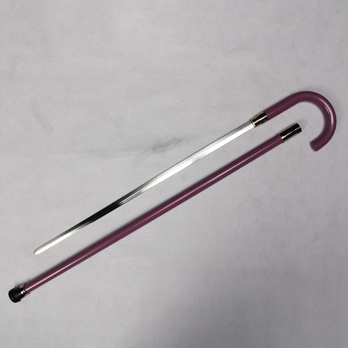Canne-épée “du Musicien Squelette” ouverte et présentée sur un fond gris clair texturé : la lame inox polie est entièrement dégainée, posée parallèlement au tube en acier laqué violet, avec la poignée en crochet et les bagues métalliques visibles ; éclairage neutre mettant en valeur les reflets du métal et la couleur profonde du fourreau.