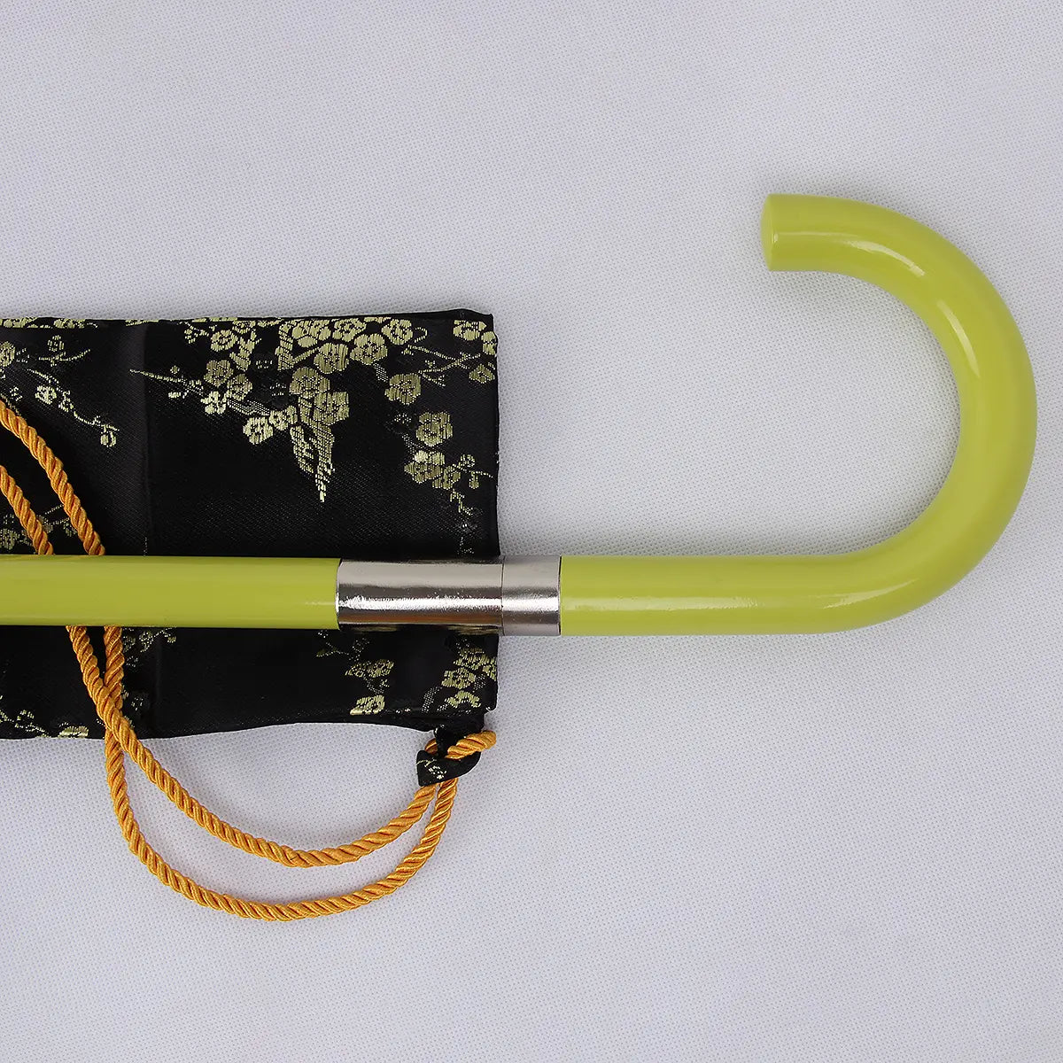 Gros plan de la poignée courbe couleur vert jade d’une canne-épée cosplay inspirée de Bleach, posée sur un tissu noir à motifs dorés ; finition laquée brillante, bague métallique argentée visible, décor éclairé sur fond gris clair neutre.