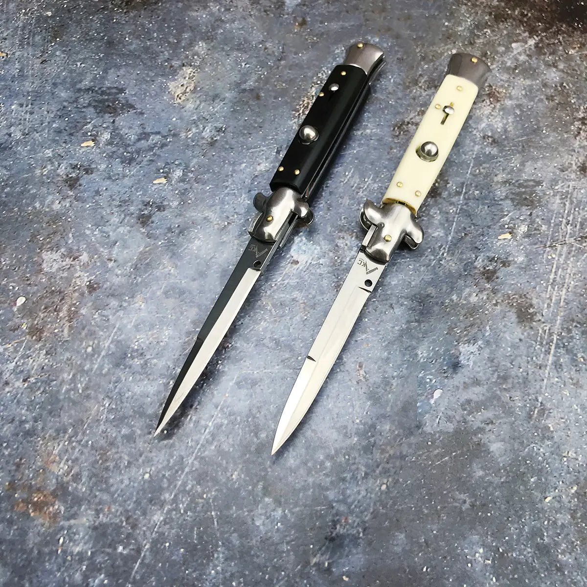 Deux couteaux automatiques italiens Stiletto ouverts, posés sur un fond bleu acier légèrement texturé. L’un possède un manche noir profond, l’autre un manche ivoire clair, tous deux dotés d’une lame en acier 440C brillante et d’un mécanisme à bouton chromé. L’éclairage froid souligne l’élégance et la précision du design – Blade Bushido.