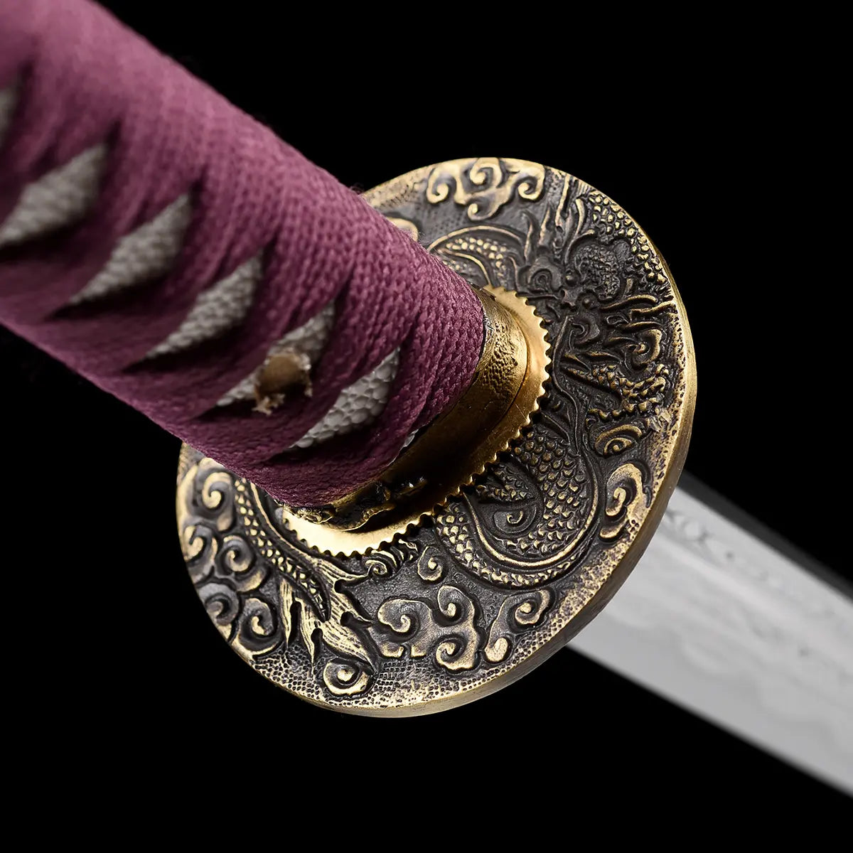 Close-up detail of the handcrafted Japanese katana Dokuryu on a black background, focusing on the round metal tsuba engraved with a Japanese dragon surrounded by traditional cloud motifs, brass habaki fitted at the base of the blade, purple tsuka wrap in tight traditional braid revealing white ray skin panels beneath, blade partially visible, minimalist studio composition with no scabbard and no stand.
