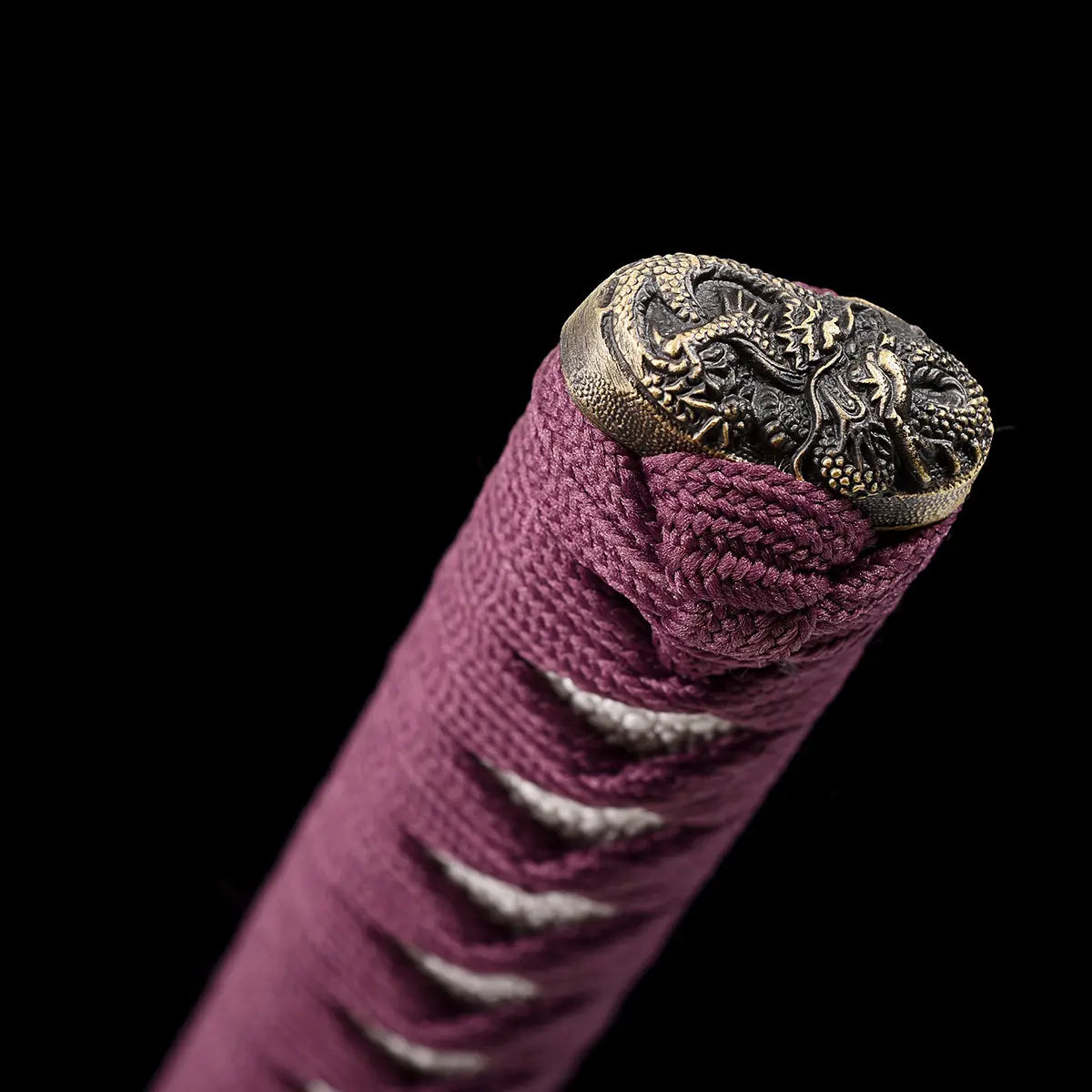 Close-up detail of the handcrafted Japanese katana Dokuryu on a black background, focusing on the kashira decorated with an engraved Japanese dragon in aged brass tone, purple tsuka wrapped in tight traditional cord with white ray skin panels partially visible, braided end knot clearly defined, minimalist studio lighting highlighting metal relief and textile texture, no blade or scabbard visible.
