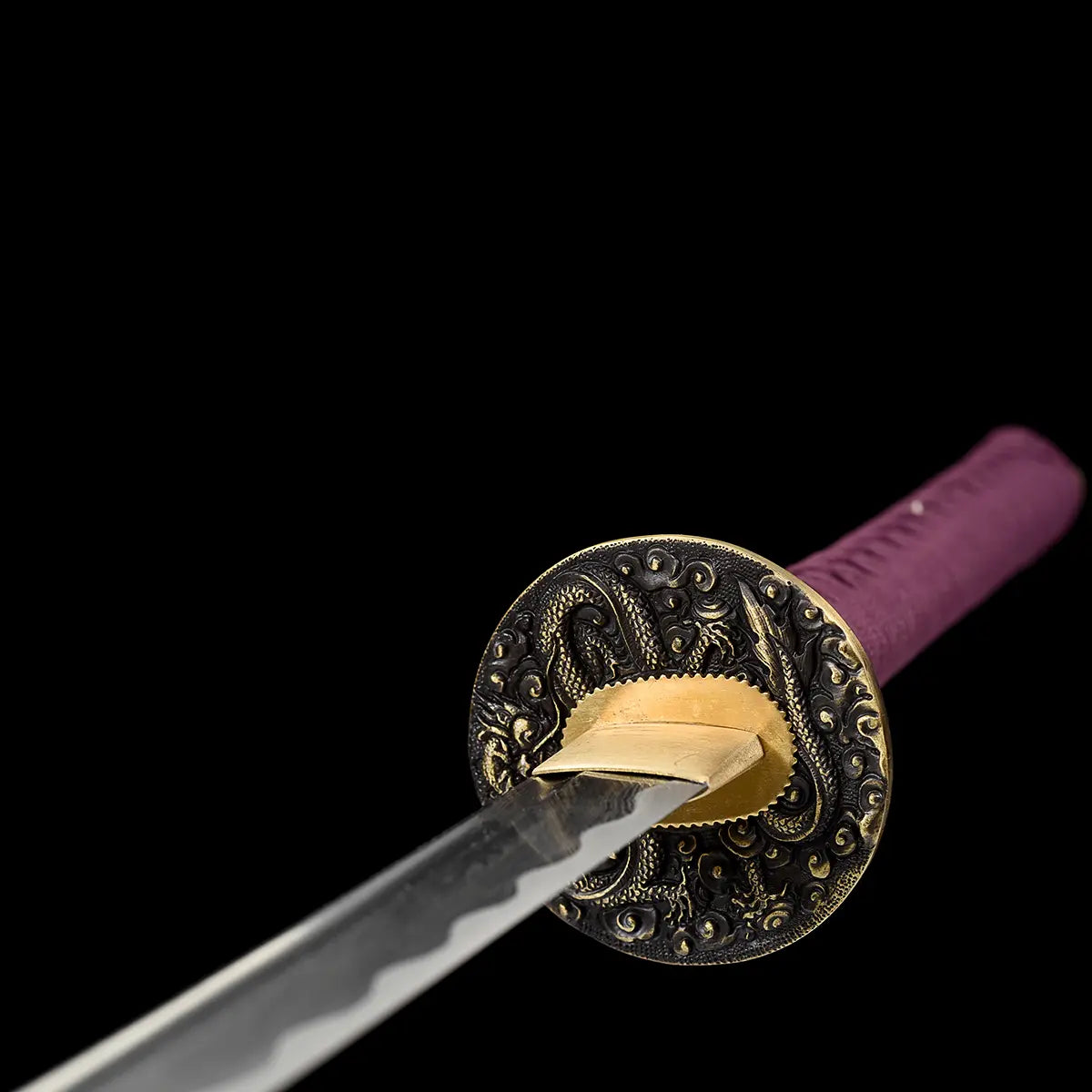 Front angle close-up of the handcrafted Japanese katana Dokuryu on a black background, showing the round metal tsuba engraved with a Japanese dragon and swirling cloud motifs, brass habaki fitted around the blade base, folded steel blade extending forward with a visible wavy hamon near the edge, purple tsuka wrap partially visible in the background, minimalist studio composition with no scabbard and no stand.
