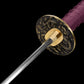 Rear angle close-up of the handcrafted Japanese katana Dokuryu on a black background, showing the back side of the round metal tsuba engraved with a Japanese dragon and cloud motifs, brass habaki tightly fitted around the blade base, folded steel blade extending forward, purple tsuka wrap partially visible, minimalist studio composition with no scabbard and no stand.
