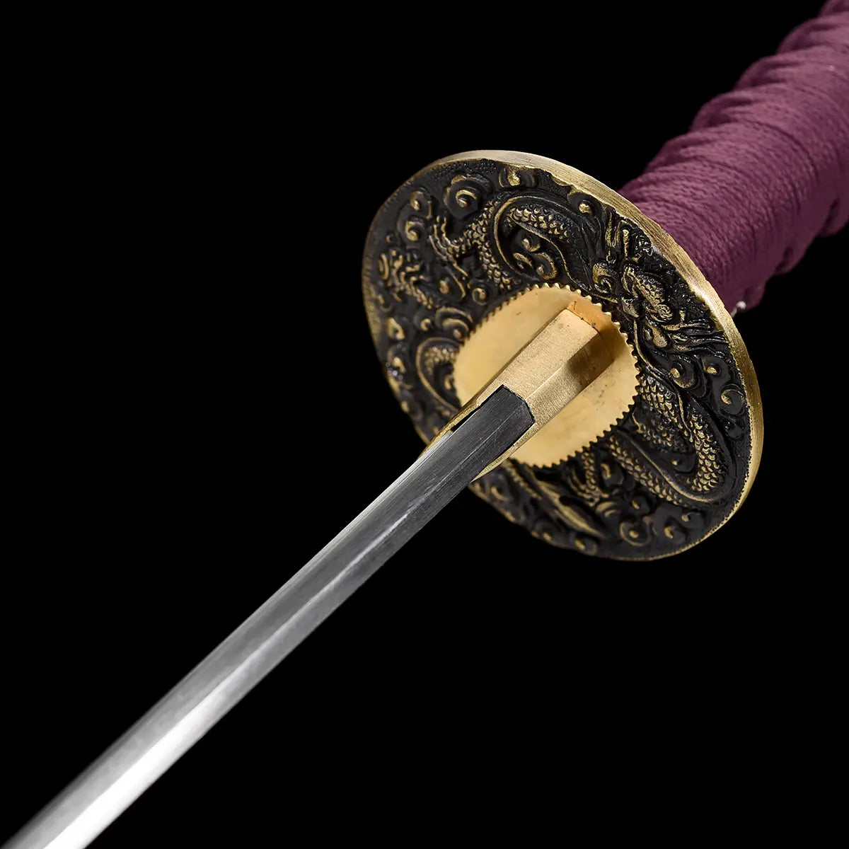 Rear angle close-up of the handcrafted Japanese katana Dokuryu on a black background, showing the back side of the round metal tsuba engraved with a Japanese dragon and cloud motifs, brass habaki tightly fitted around the blade base, folded steel blade extending forward, purple tsuka wrap partially visible, minimalist studio composition with no scabbard and no stand.

