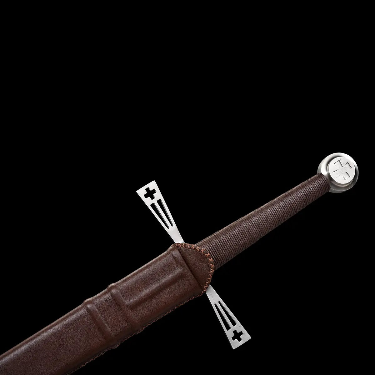 Gros plan de l’épée longue médiévale Crux Bellator partiellement rangée dans son fourreau en cuir brun cousu, montrant la garde en croix ajourée en acier brillant, la poignée gainée de cuir foncé et le pommeau rond métallique gravé, photographiés sur fond noir pour mettre en valeur les textures et les finitions.