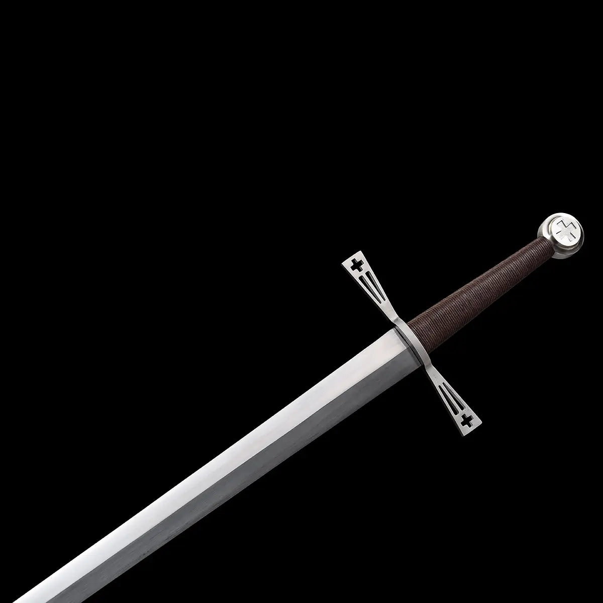 Vue rapprochée de l’épée longue médiévale Crux Bellator montrant la garde en croix ajourée en acier brillant, la poignée gainée de cuir brun enroulé, le pommeau rond métallique gravé ainsi que la base de la lame en acier carbone 1060 polie, le tout photographié sur fond noir uniforme pour accentuer les détails et les matériaux.