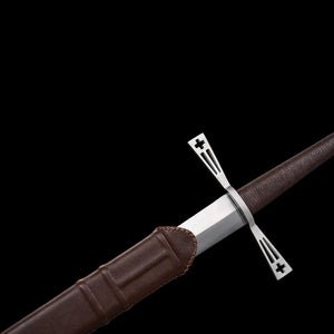 Gros plan de l’épée longue médiévale Crux Bellator partiellement insérée dans son fourreau en cuir brun, montrant la garde en croix ajourée en acier brillant, la base de la lame en acier carbone 1060 polie et la texture cousue du cuir, photographiés sur fond noir uniforme pour mettre en valeur les matériaux et les finitions.