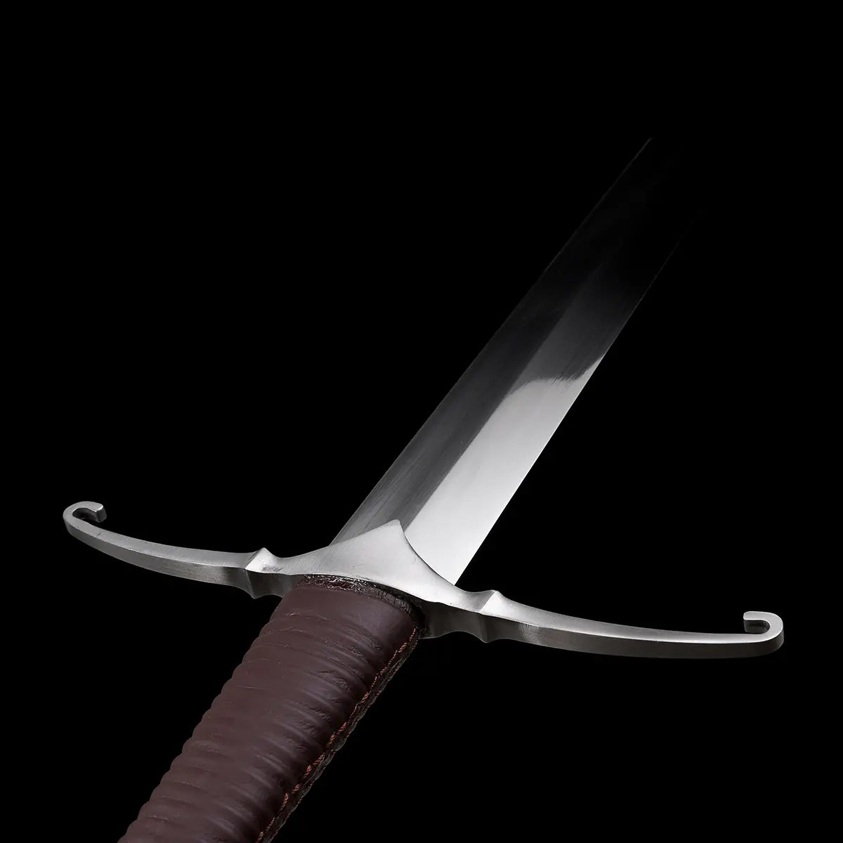 Gros plan immersif sur la garde courbée de l’épée longue médiévale Castelar, photographiée sur fond noir. On distingue la lame droite en acier 1060 polie miroir, les quillons incurvés vers l’avant, ainsi que la poignée gainée de cuir brun foncé avec couture visible. L’image met en valeur le style authentique du longsword médiéval, la qualité du polissage et la jonction garde-lame. Optimisée pour les recherches épée médiévale, garde courbée, longsword 1060, détail poignée cuir.