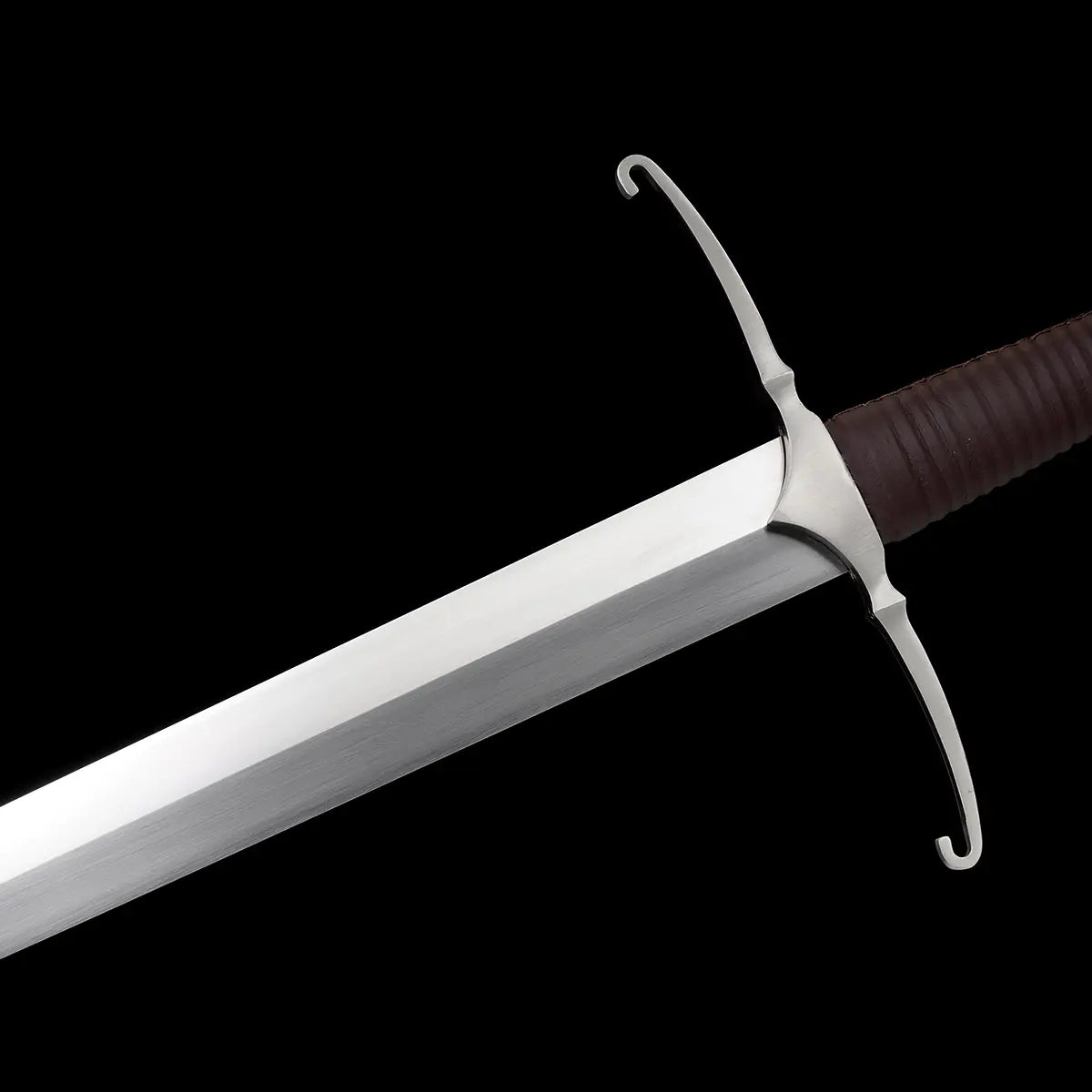 Gros plan sur la garde en croix de l’épée longue médiévale Castelar, photographiée sur fond noir. On distingue la lame droite en acier 1060 polie, les quillons courbés argentés, ainsi que la poignée gainée de cuir brun foncé. L’image met en valeur les finitions métalliques, la jonction entre la lame et la garde, et le style authentique de longsword médiéval. Idéal pour les mots-clés épée longue médiévale, garde en croix, longsword acier 1060, détail forge.