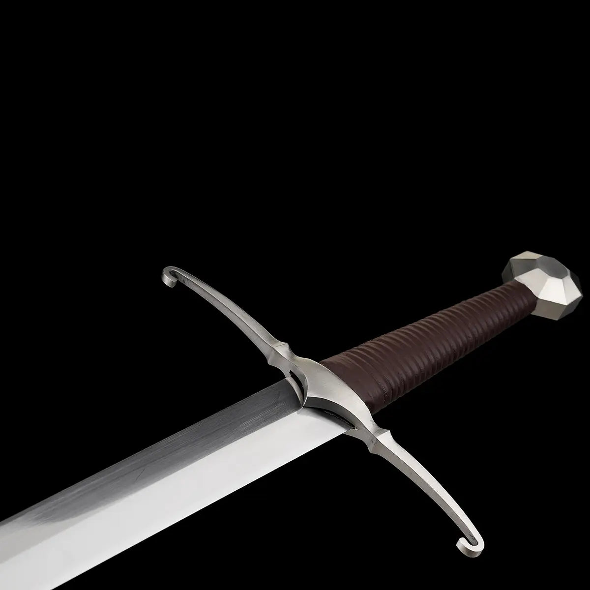 Gros plan sur la poignée et le pommeau facetté de l’épée longue médiévale Castelar, sur fond noir uni. La lame en acier 1060 apparaît partiellement, avec sa surface polie et son biseau net. La garde en croix courbée est bien visible, ainsi que la poignée gainée de cuir brun foncé. L’image met en avant les finitions métalliques, la géométrie du pommeau et le style authentique d’un longsword médiéval. Optimisée pour les mots-clés épée longue médiévale, détail pommeau, poignée cuir, longsword acier 1060.