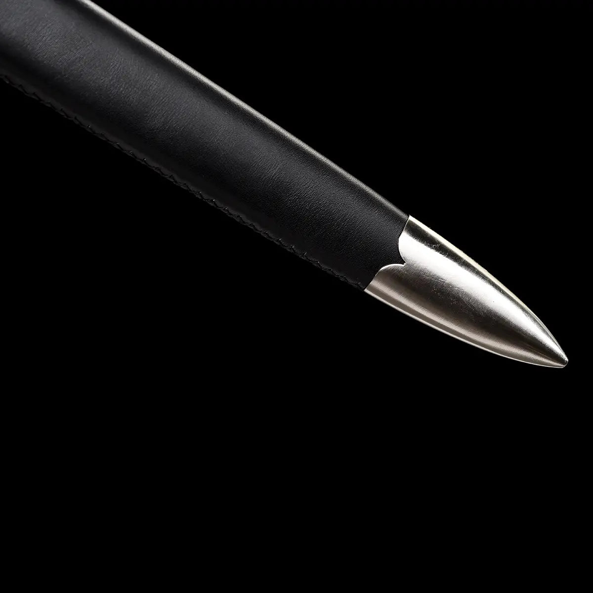 Gros plan sur l’extrémité du fourreau de l’épée longue médiévale Corvus, embout métallique poli protégeant la pointe, fourreau en bois gainé de cuir noir avec coutures visibles, finition robuste et fonctionnelle, photographié sur fond noir profond pour une ambiance sobre, martiale et haut de gamme.