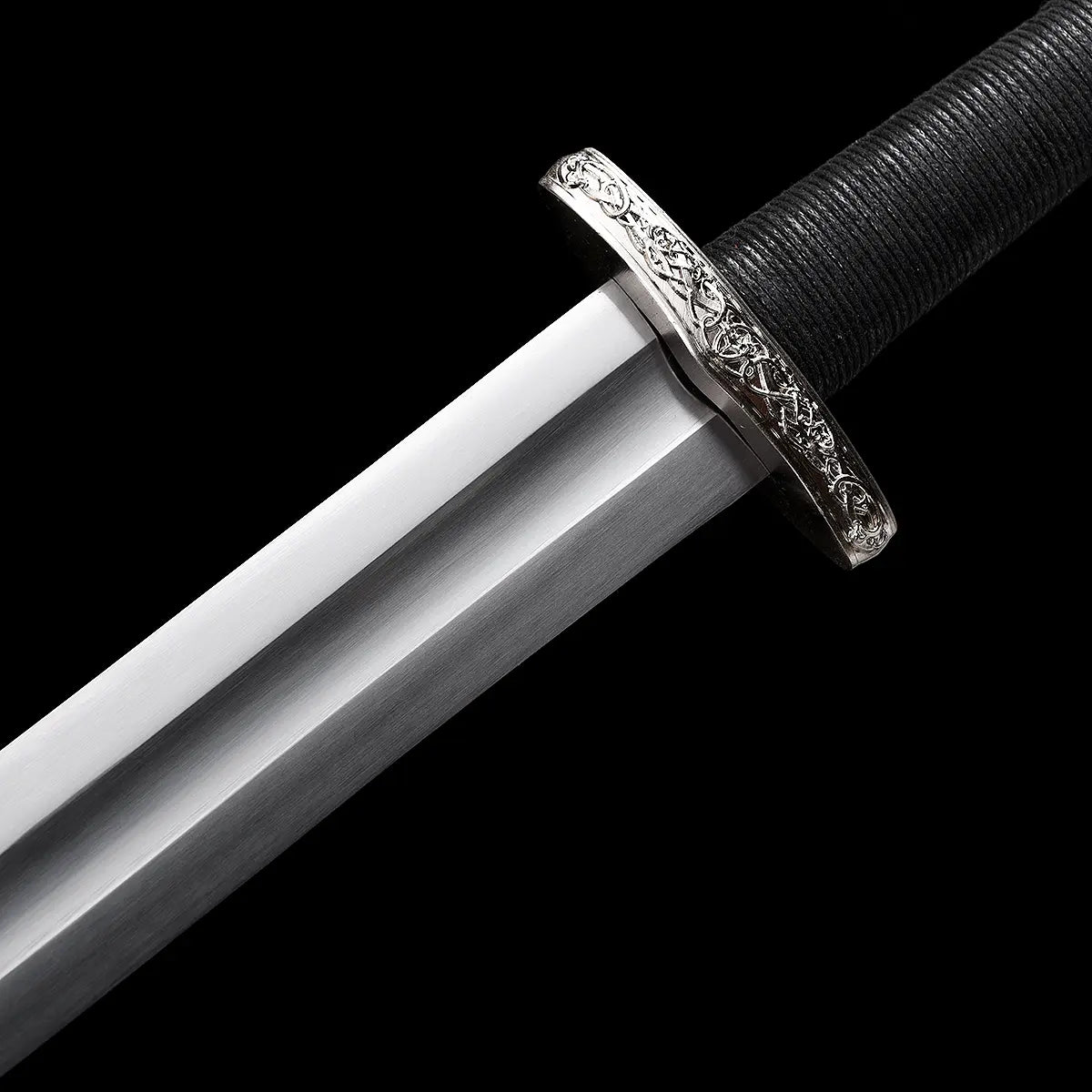 Détail de la garde et de la base de lame de l’épée longue médiévale Ravenna sur fond noir uniforme. Garde en croix courte en métal argenté finement texturé, lame droite en acier 1060 avec polissage satiné et poignée gainée de cuir noir offrant une excellente ergonomie. Finition fonctionnelle et équilibrée, adaptée à la pratique AMHE / HEMA, à l’entraînement ou à la collection médiévale.