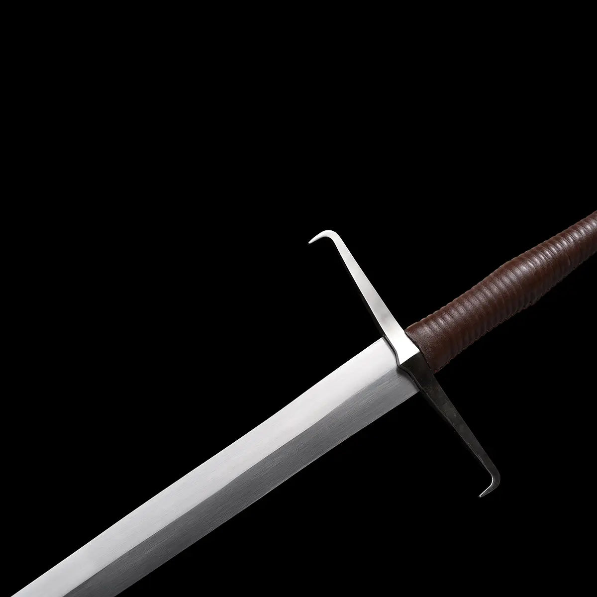 Gros plan de l’épée longue médiévale Vesper montrant la garde en croix droite, les quillons légèrement recourbés, la base de la lame en acier 1060 polie et la poignée gainée de cuir brun, le tout mis en valeur par un fond noir contrasté — longsword main-et-demi, épée médiévale 1060, finition cuir brun.