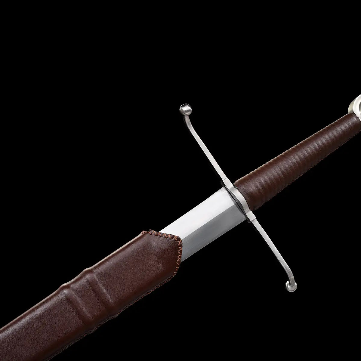 Épée longue médiévale Valmont en acier 1060 photographiée sur fond noir, vue rapprochée de la garde en croix et de la base de lame partiellement engagée dans le fourreau en cuir brun cousu main, poignée gainée de cuir brun, finition sobre et martiale, style longsword main-et-demi pour collection, décoration ou pratique HEMA légère
