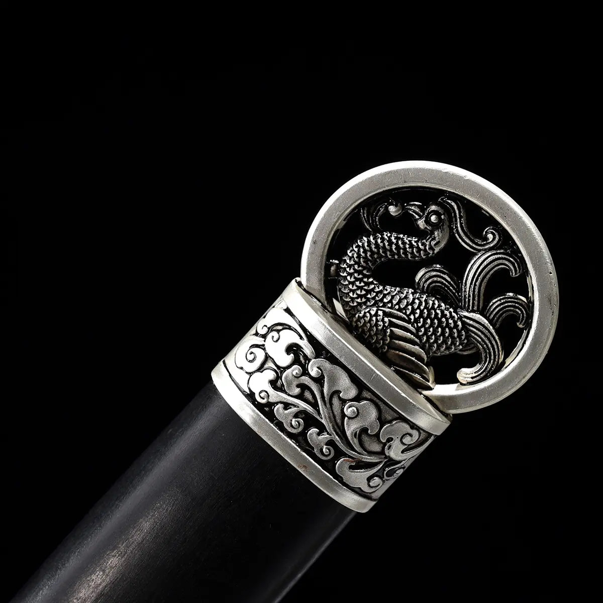 Gros plan sur le pommeau argenté décoré d’un phénix de l’épée chinoise Jian FENG YIN, photographié sur fond noir. Le Jian chinois présente une poignée noire, des garnitures métalliques gravées et un pommeau circulaire travaillé avec motif d’oiseau mythique.