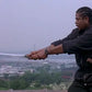 Side view of a Japanese shirasaya katana held with both hands by a man dressed in black, curved steel blade fully drawn from its wooden scabbard, light gray sky background with an industrial city landscape in the distance, natural outdoor lighting, minimalist wooden handle and saya with smooth finish, no guard visible, sharp tip and continuous curvature of the blade clearly defined
