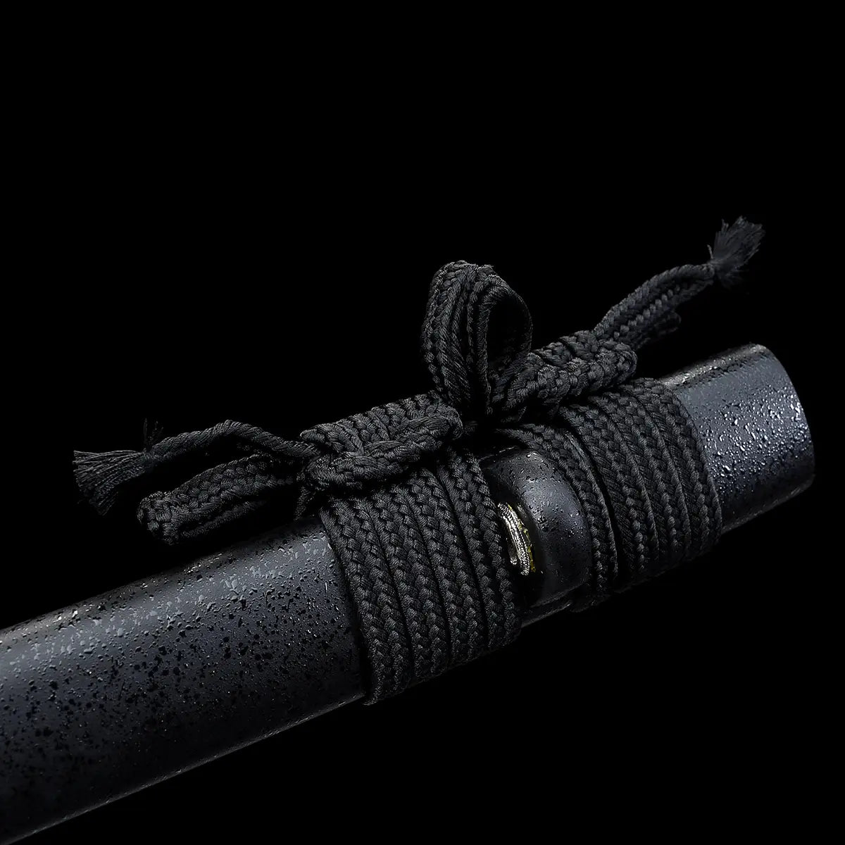 Détail du fourreau saya du katana japonais artisanal Kazekiri, saya en bois dur avec finition laquée noire mate texturée, cordon sageo noir épais soigneusement noué autour du fourreau, surface légèrement granuleuse visible, fond noir uniforme mettant en valeur la sobriété et la finition du fourreau
