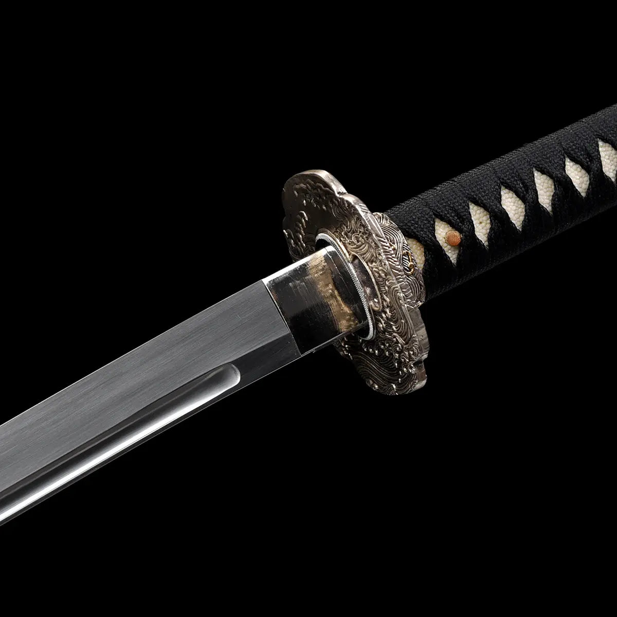 Gros plan sur la base de la lame du katana japonais artisanal Kazekiri, montrant le habaki ajusté et la tsuba finement sculptée, photographie sur fond noir profond, lame en acier poli avec rainure visible près du dos, habaki métallique brillant aux reflets argentés, tsuba en métal décoré de motifs ondulés en relief, tsuka partiellement visible avec tressage ito noir et losanges de samegawa claire, absence de fourreau, mise au point précise sur les détails d’assemblage et de finition
