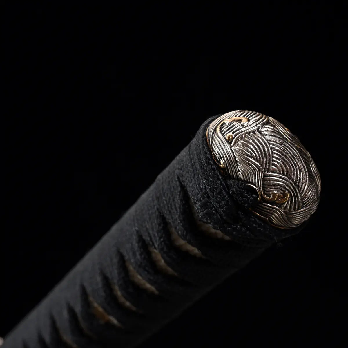 Gros plan sur l’extrémité du manche tsuka du katana japonais artisanal Kazekiri, kashira en métal finement sculpté avec motifs ondulés et détails dorés, tressage ito noir serré et régulier visible autour du manche, texture textile marquée, absence de fourreau sur l’image, fond noir profond mettant en valeur les reliefs, les gravures et les finitions métalliques

