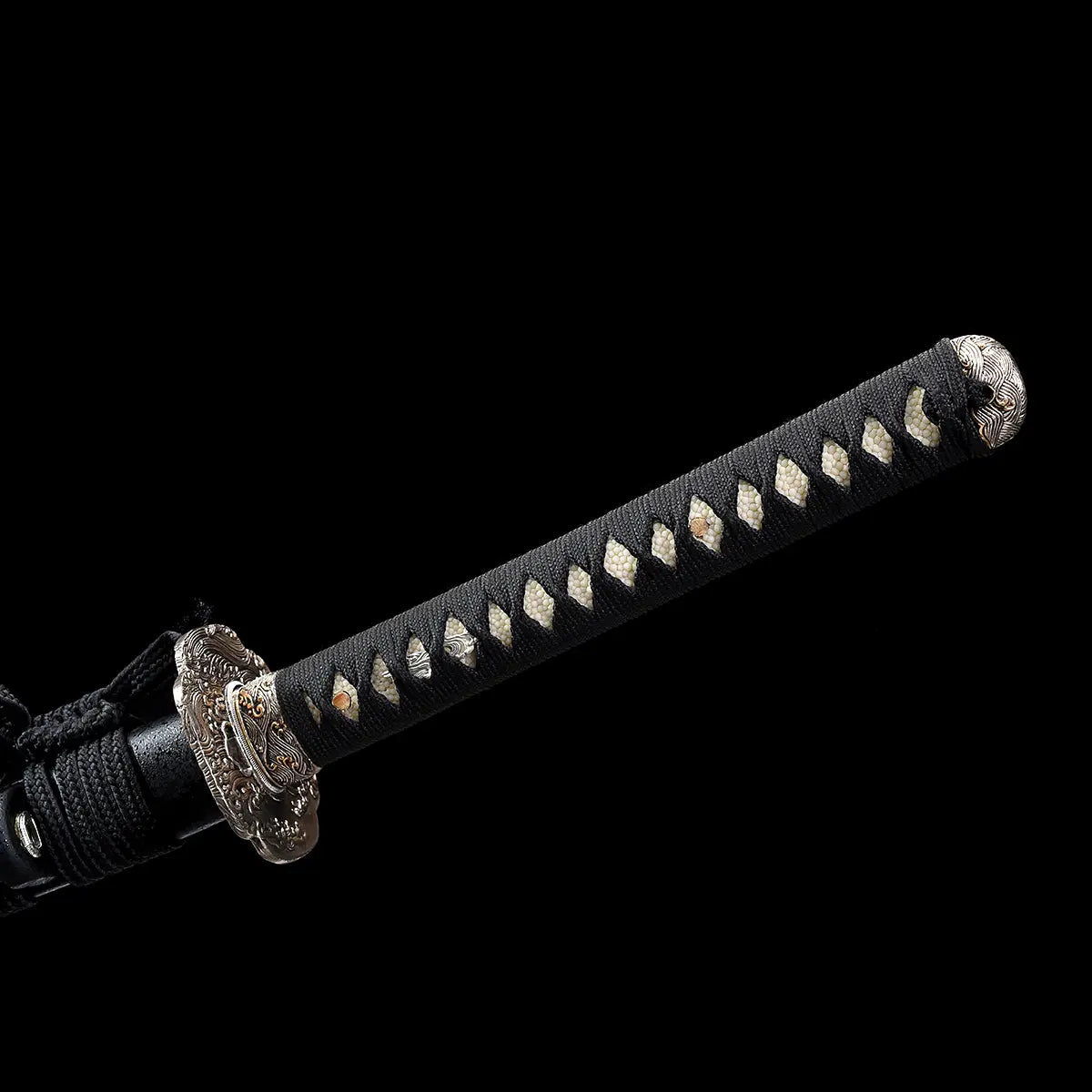 Gros plan sur le manche tsuka du katana japonais artisanal Kazekiri, manche recouvert d’un tressage ito noir serré laissant apparaître la peau de raie blanche samegawa, motifs losangés réguliers visibles, extrémité kashira métallique décorée, garde tsuba en laiton partiellement visible, fond noir uniforme mettant en valeur la texture et la qualité de l’assemblage
