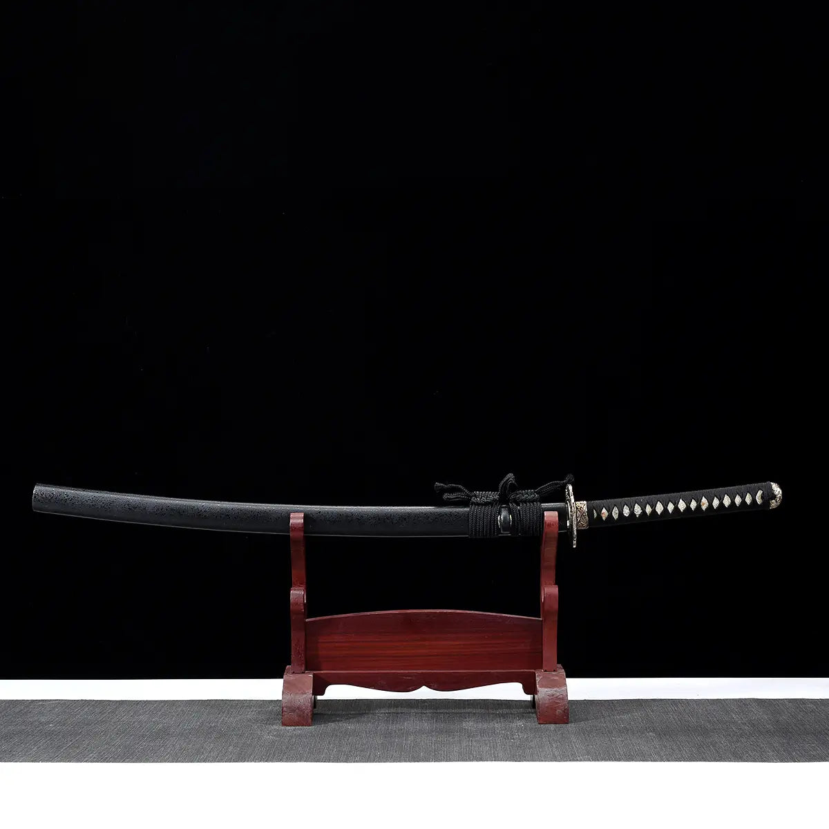 Japanese artisanal katana Kazekiri displayed horizontally on a red wooden stand, matte black lacquered saya with black sageo cord, black tsuka wrapped with black ito over white ray skin, silver-toned tsuba, dark black background with neutral grey surface, full sword shown in profile without the blade drawn
