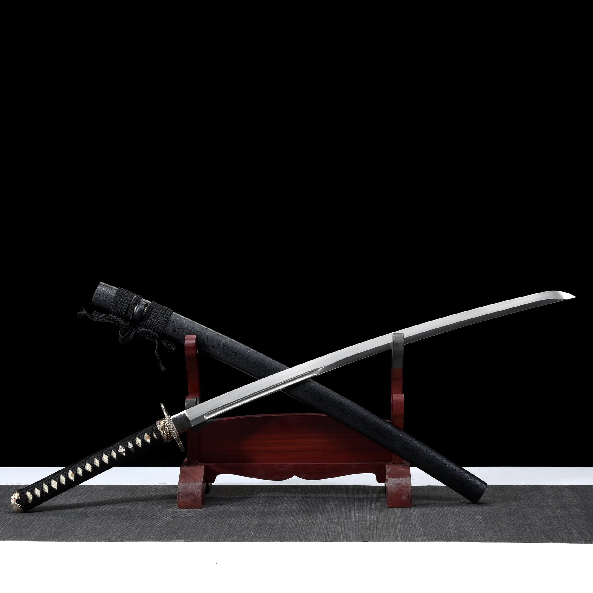 Katana japonais artisanal Kazekiri présenté dégainé en position croisée sur un support en bois rouge, lame en acier carbone 1060 visible avec géométrie unokubi-zukuri et courbure marquée, fourreau saya noir laqué mat disposé en diagonale, manche tsuka noir tressé en ito noir sur peau de raie blanche, garde tsuba métallique détaillée, fond noir profond avec surface gris neutre
