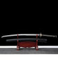 Katana japonais artisanal Kazekiri présenté dégainé sur un support en bois rouge, lame en acier carbone 1060 visible avec géométrie unokubi-zukuri, fourreau saya noir laqué mat placé en dessous, manche tsuka noir avec tressage ito noir sur peau de raie blanche, garde tsuba métallique décorée, fond noir profond avec surface gris neutre

