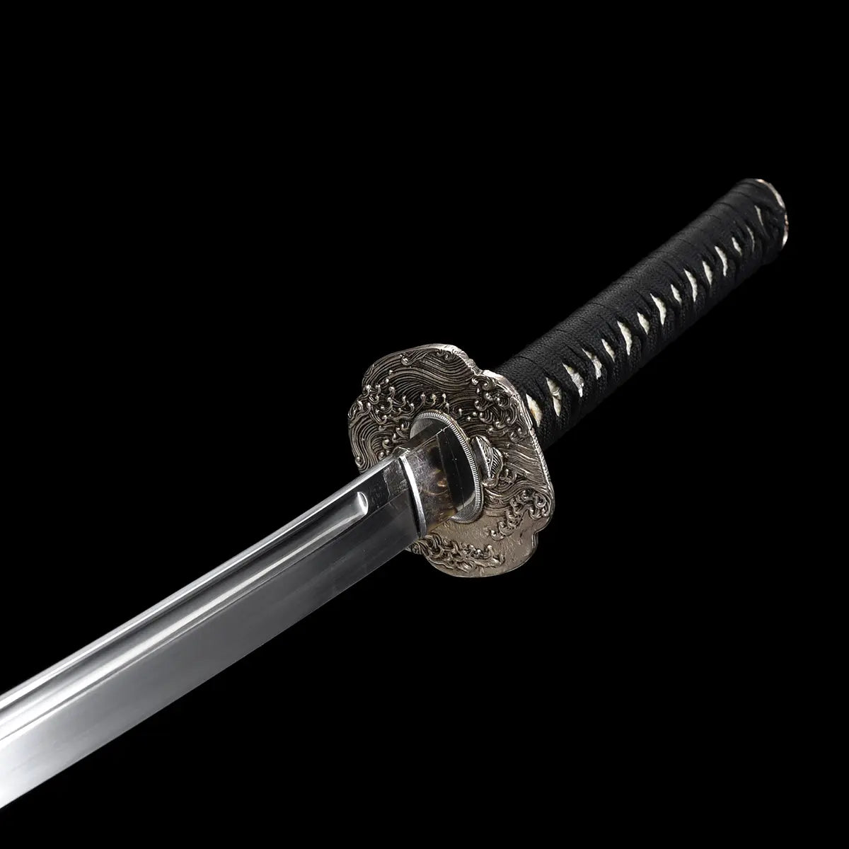 Vue rapprochée du katana japonais artisanal Kazekiri lame dégainée, montrant la garde tsuba en laiton finement décorée, le habaki ajusté et la base de la lame en acier carbone 1060 avec gorge bo-hi visible, manche tsuka noir tressé en ito noir laissant apparaître la peau de raie blanche, fond noir uniforme mettant en valeur les lignes et la finition du sabre
