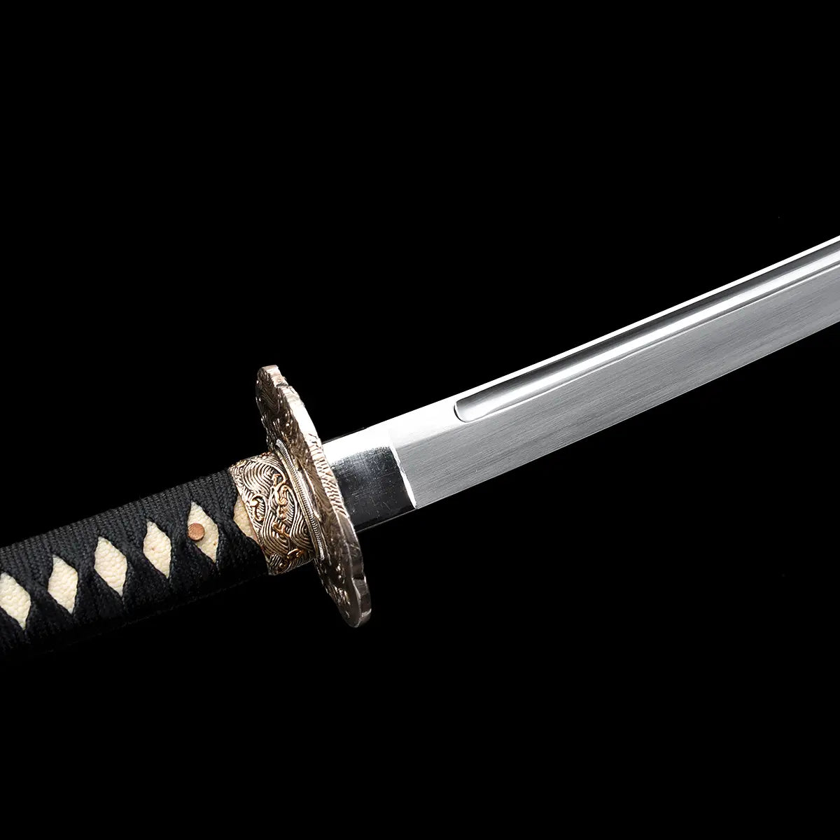 Détail de la base de la lame du katana japonais artisanal Kazekiri montrant le habaki ajusté, le début de la gorge bo-hi et la finition satinée de l’acier carbone 1060, garde tsuba en laiton finement décorée visible en arrière-plan, manche tsuka noir tressé en ito noir sur peau de raie blanche, fond noir uniforme mettant en valeur les lignes et la précision de l’assemblage
