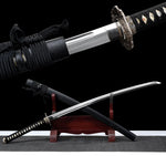 Détail rapproché du katana japonais artisanal Kazekiri montrant le habaki en laiton, la base de la lame en acier carbone 1060 avec géométrie unokubi-zukuri et gorge visible, la garde tsuba finement décorée, manche tsuka noir tressé en ito noir sur peau de raie blanche, fourreau saya noir laqué mat avec sageo noir, fond noir profond avec vue secondaire du sabre dégainé sur support en bois rouge
