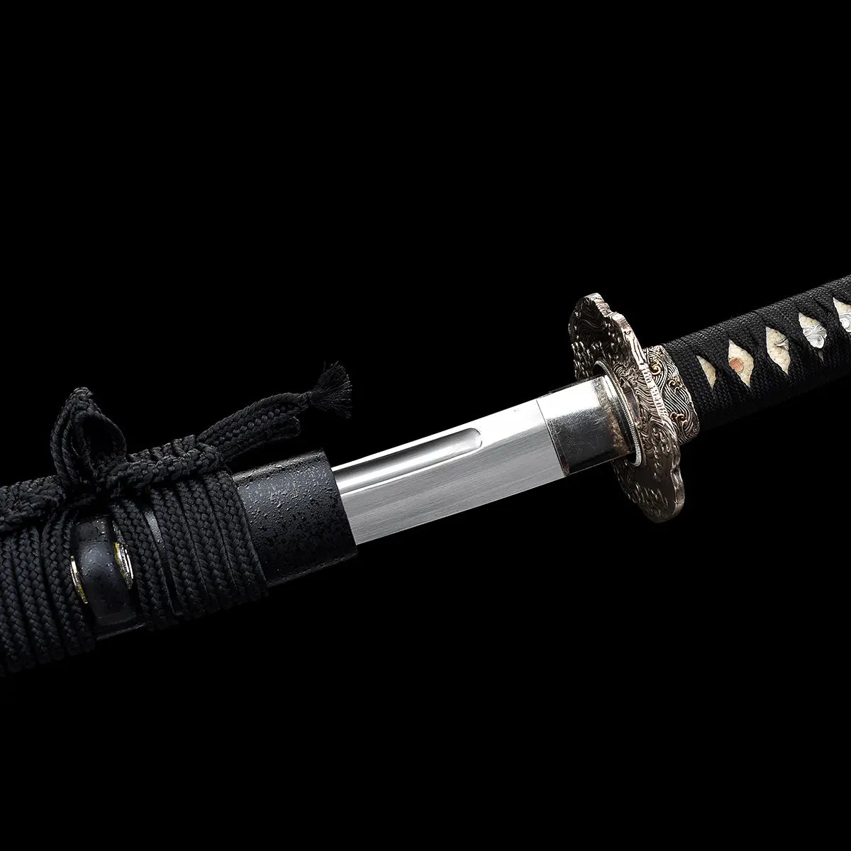 Détail de la jonction entre le fourreau et la lame du katana japonais artisanal Kazekiri, saya noir laqué mat avec sageo noir noué, habaki métallique ajusté à la base de la lame en acier carbone 1060 avec gorge bo-hi visible, manche tsuka noir tressé en ito noir laissant apparaître la peau de raie blanche, fond noir uniforme mettant en valeur la précision de l’assemblage
