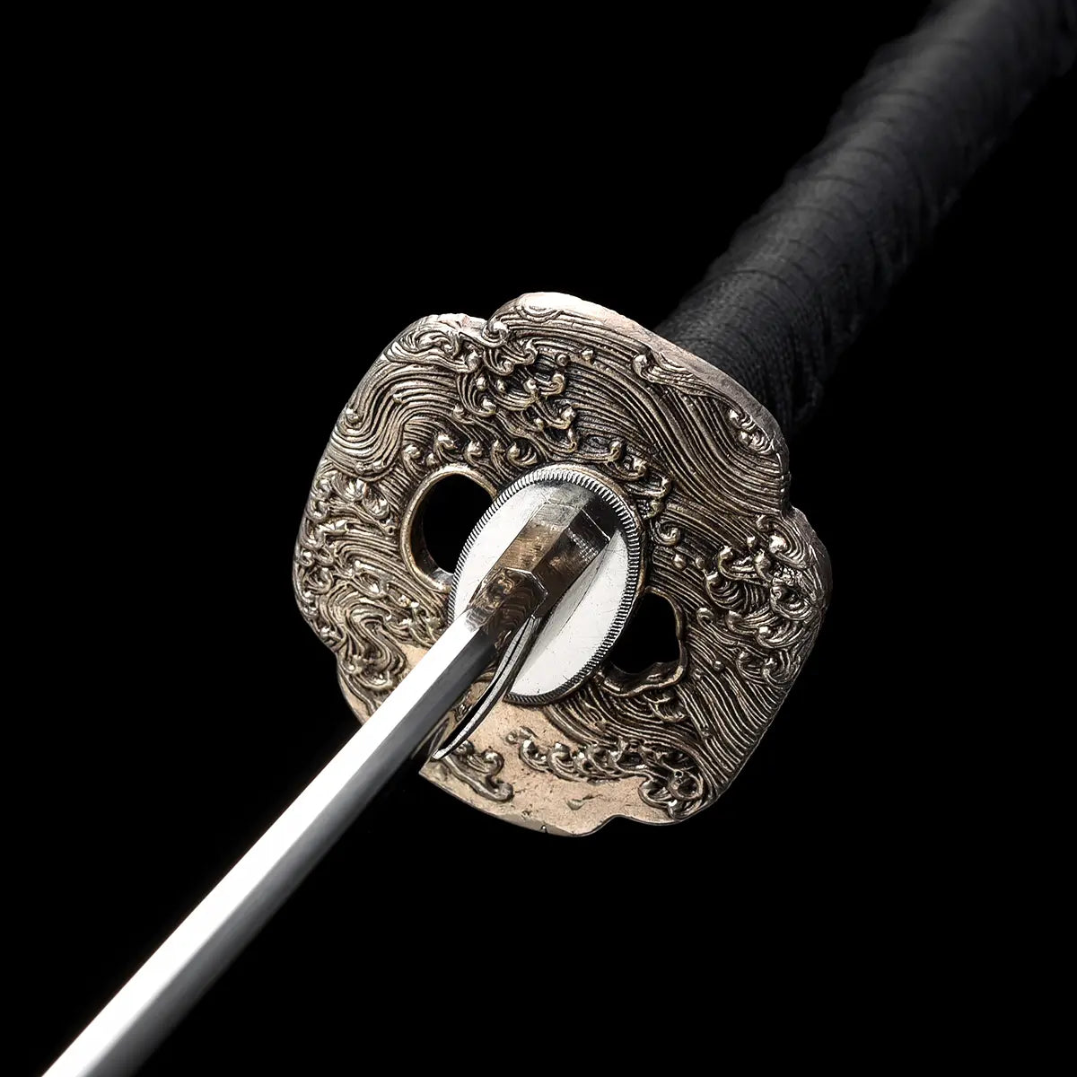 Gros plan frontal sur la garde tsuba du katana japonais artisanal Kazekiri, tsuba en laiton richement sculptée avec motifs ondulés en relief, habaki métallique ajusté autour de la base de la lame en acier carbone 1060, lame dégainée visible au centre, manche tsuka noir tressé en ito noir, fond noir profond mettant en valeur les détails et la précision de la fabrication
