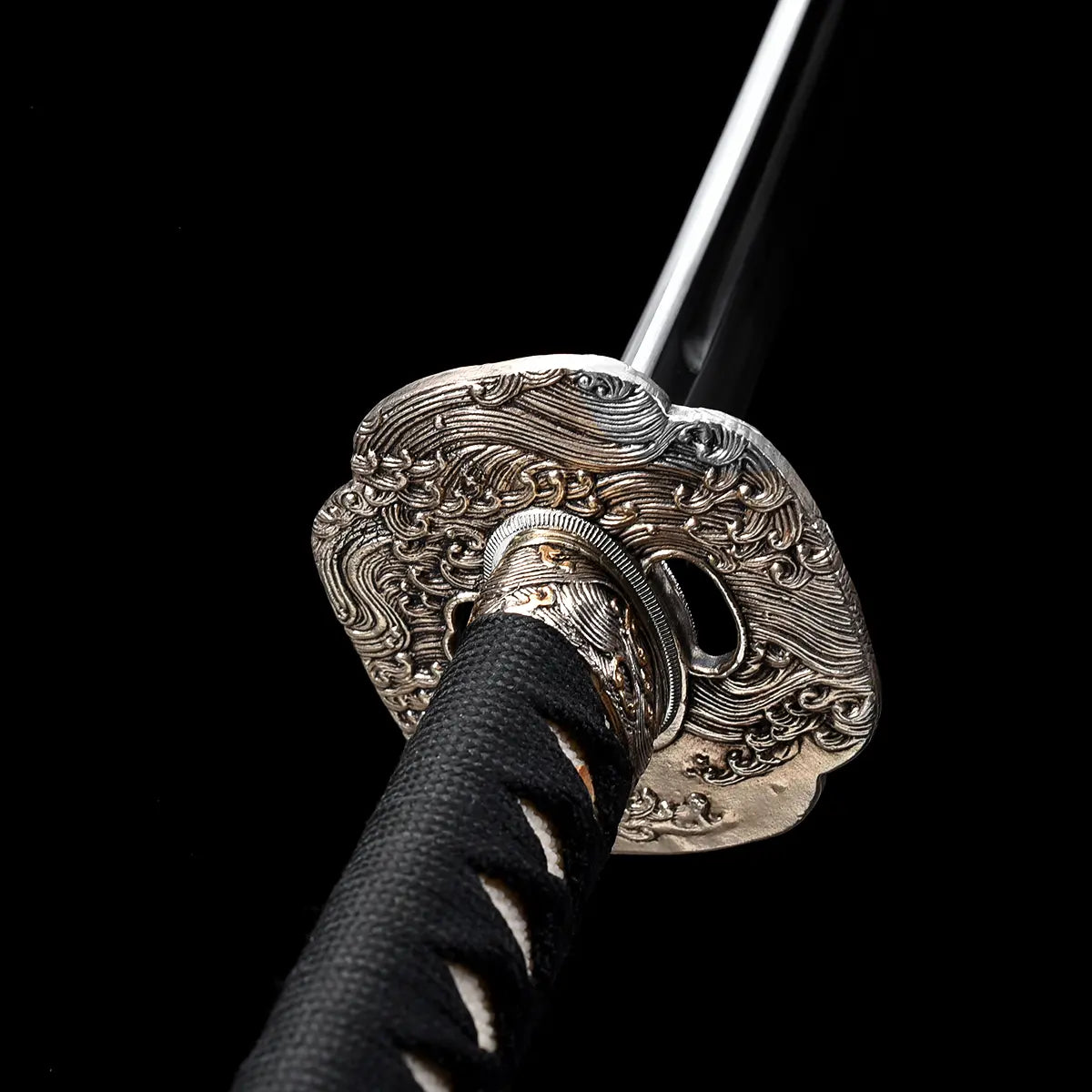 Gros plan sur la garde tsuba du katana japonais artisanal Kazekiri, tsuba en laiton finement sculptée avec motifs ondulés, habaki métallique ajusté à la base de la lame en acier carbone 1060, manche tsuka gainé de cuir noir avec tressage ito noir laissant apparaître la peau de raie blanche, lame dégainée visible en arrière-plan, fond noir profond mettant en valeur les détails de fabrication
