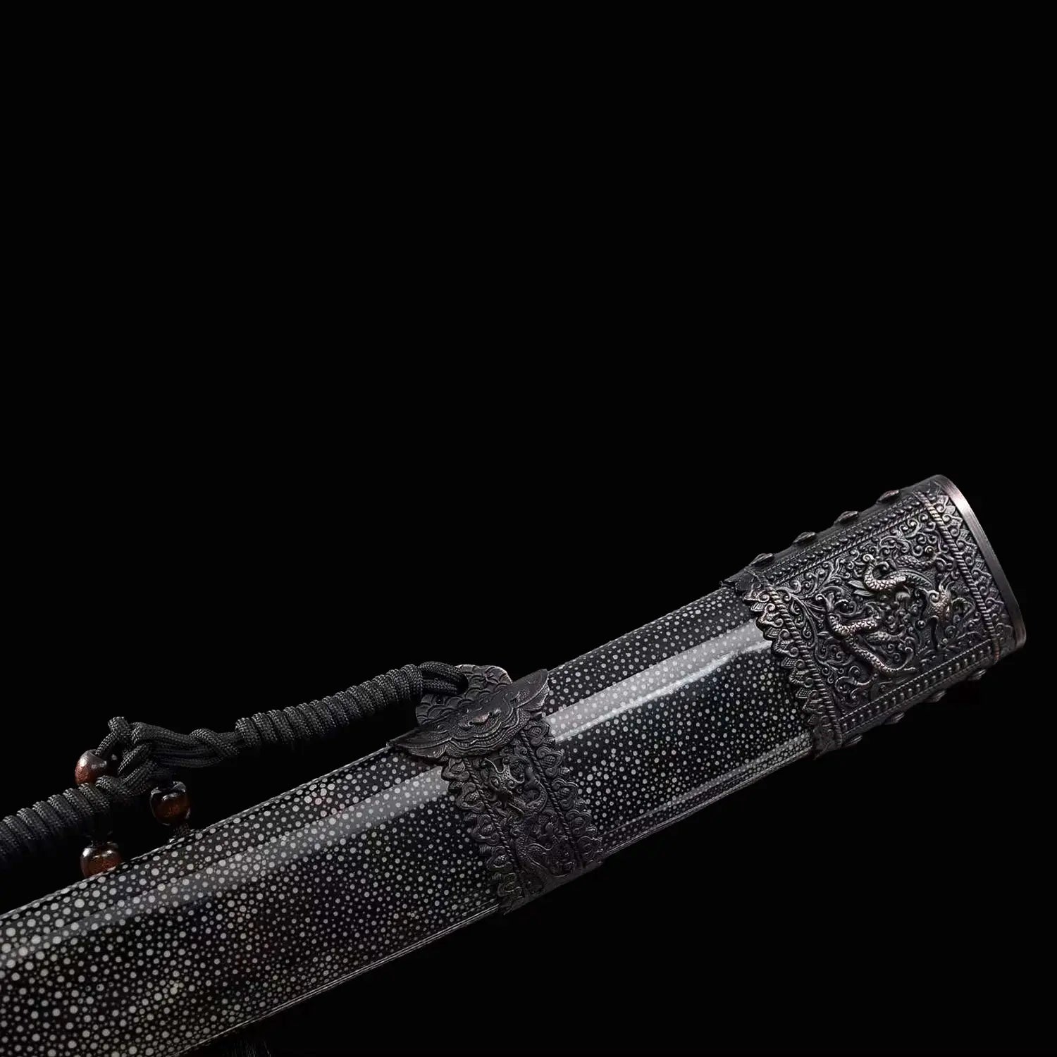 Gros plan du fourreau noir du sabre chinois Dao KANGXI LONG, avec texture mouchetée, garnitures métalliques sombres gravées, motif de dragon visible, cordon noir et perles décoratives sur fond noir.