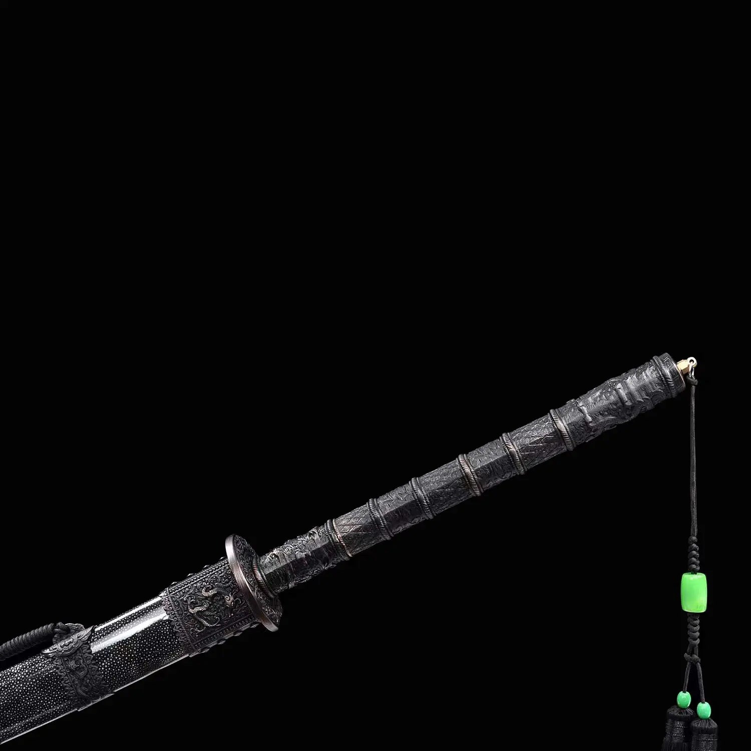 Gros plan de la poignée noire du sabre chinois Dao KANGXI LONG, avec garde ronde, garnitures gravées, cordon décoratif, perles vertes et extrémité du fourreau noir moucheté sur fond noir.