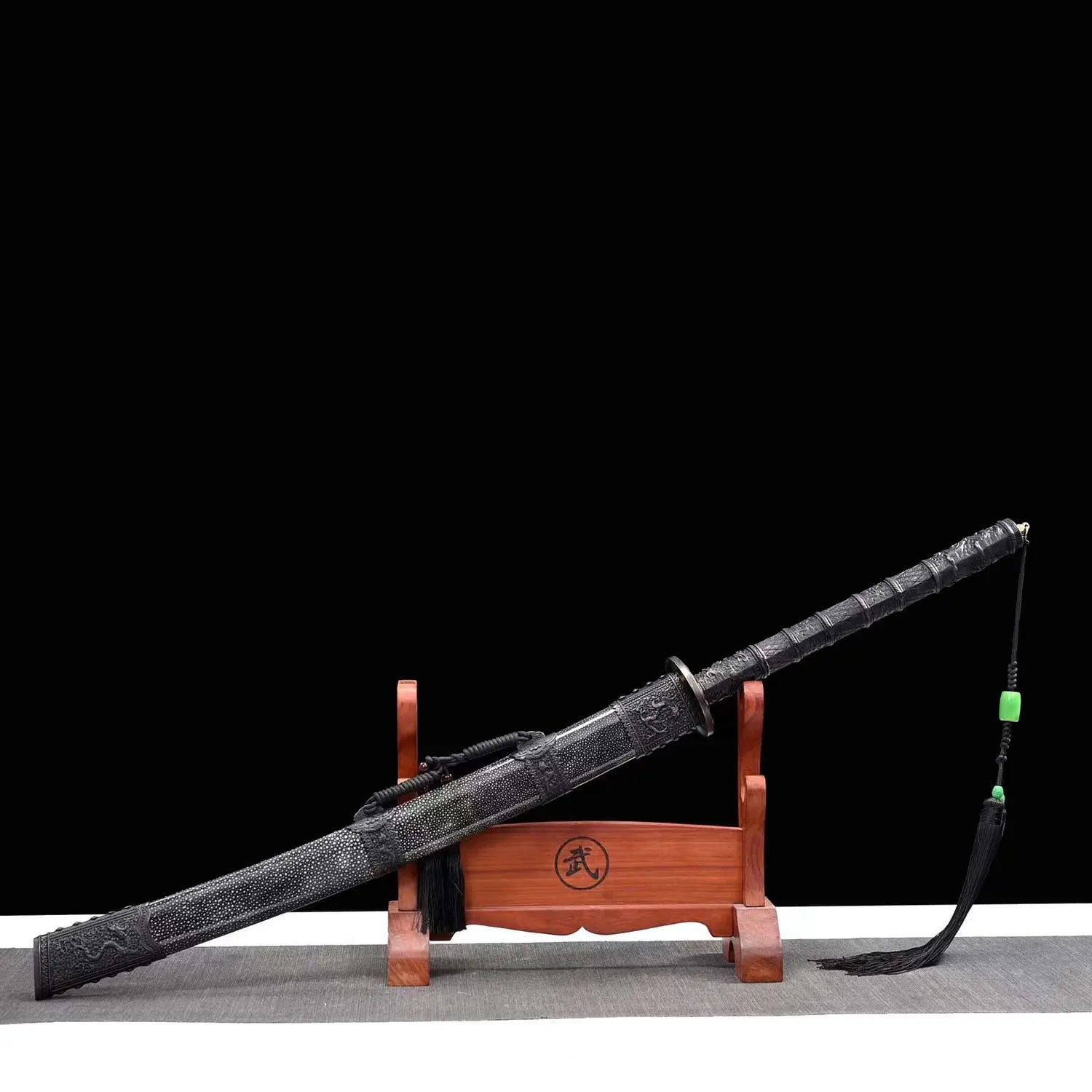 Sabre chinois Dao KANGXI LONG présenté dans son fourreau noir texturé sur un support en bois, avec poignée sombre, garde ronde, ornements gravés, pompon noir et perles vertes sur fond noir.