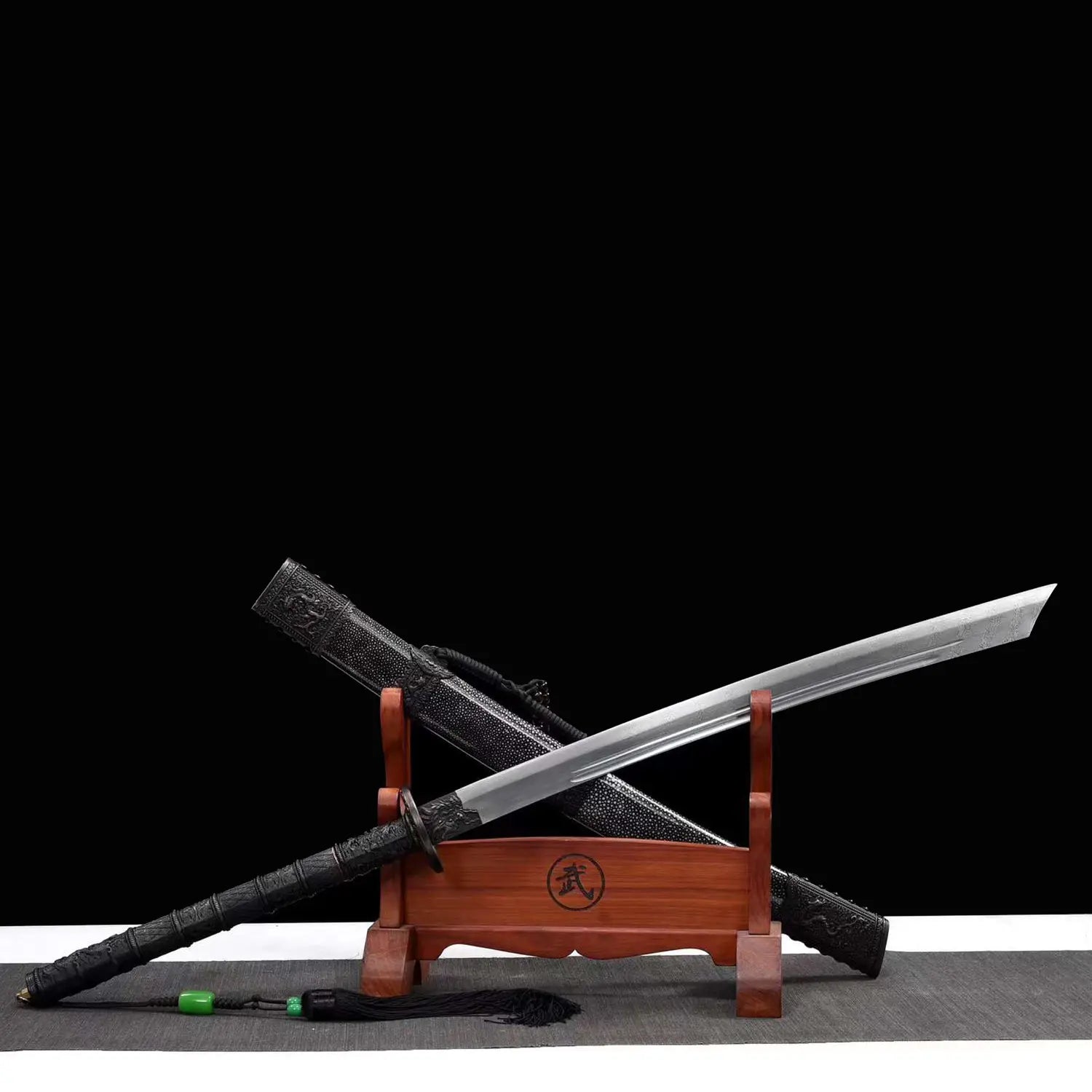 Sabre chinois Dao KANGXI LONG présenté en diagonale sur un support en bois, avec lame courbe argentée à double gorge, poignée noire, garde ronde, fourreau noir texturé et pompon avec perles vertes sur fond noir.
