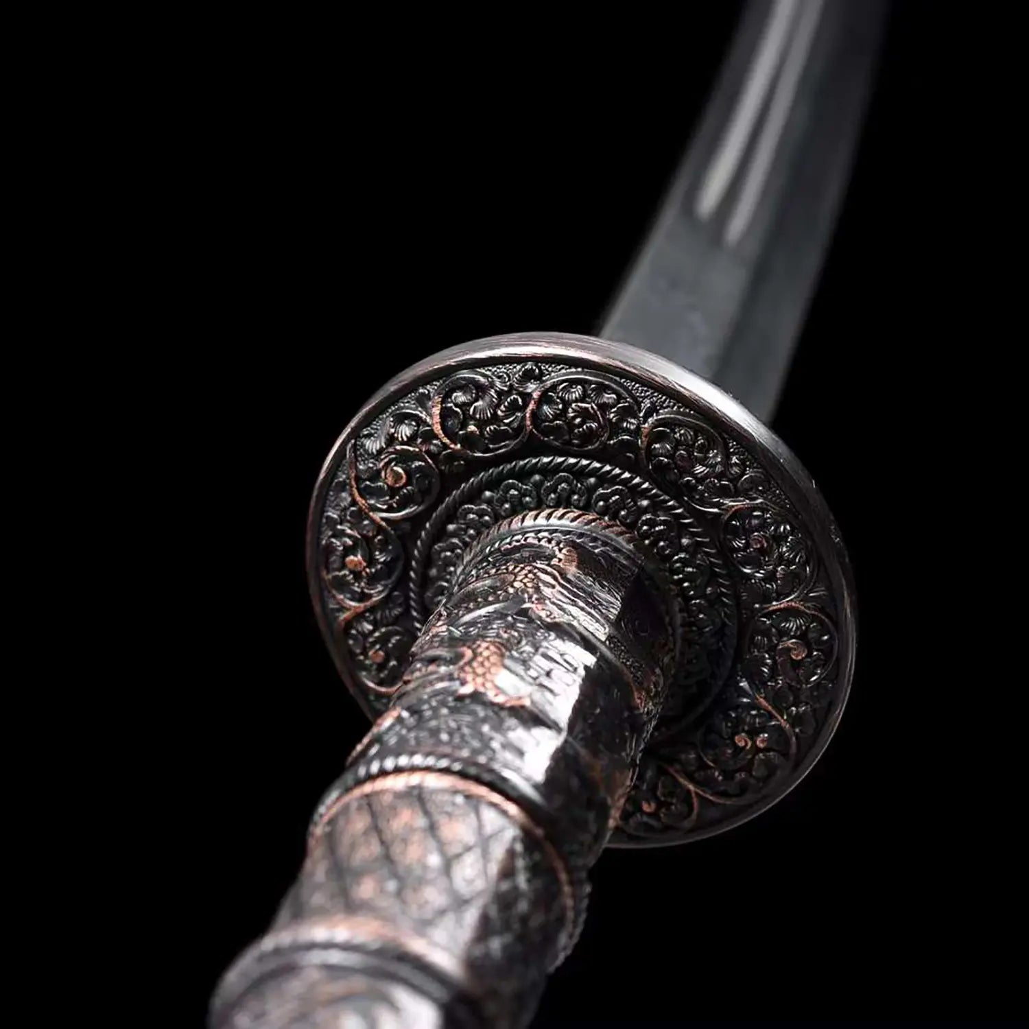 Gros plan du sabre chinois Dao KANGXI LONG avec garde ronde gravée, poignée noire texturée aux motifs métalliques sombres et lame courbe floutée en arrière-plan sur fond noir.